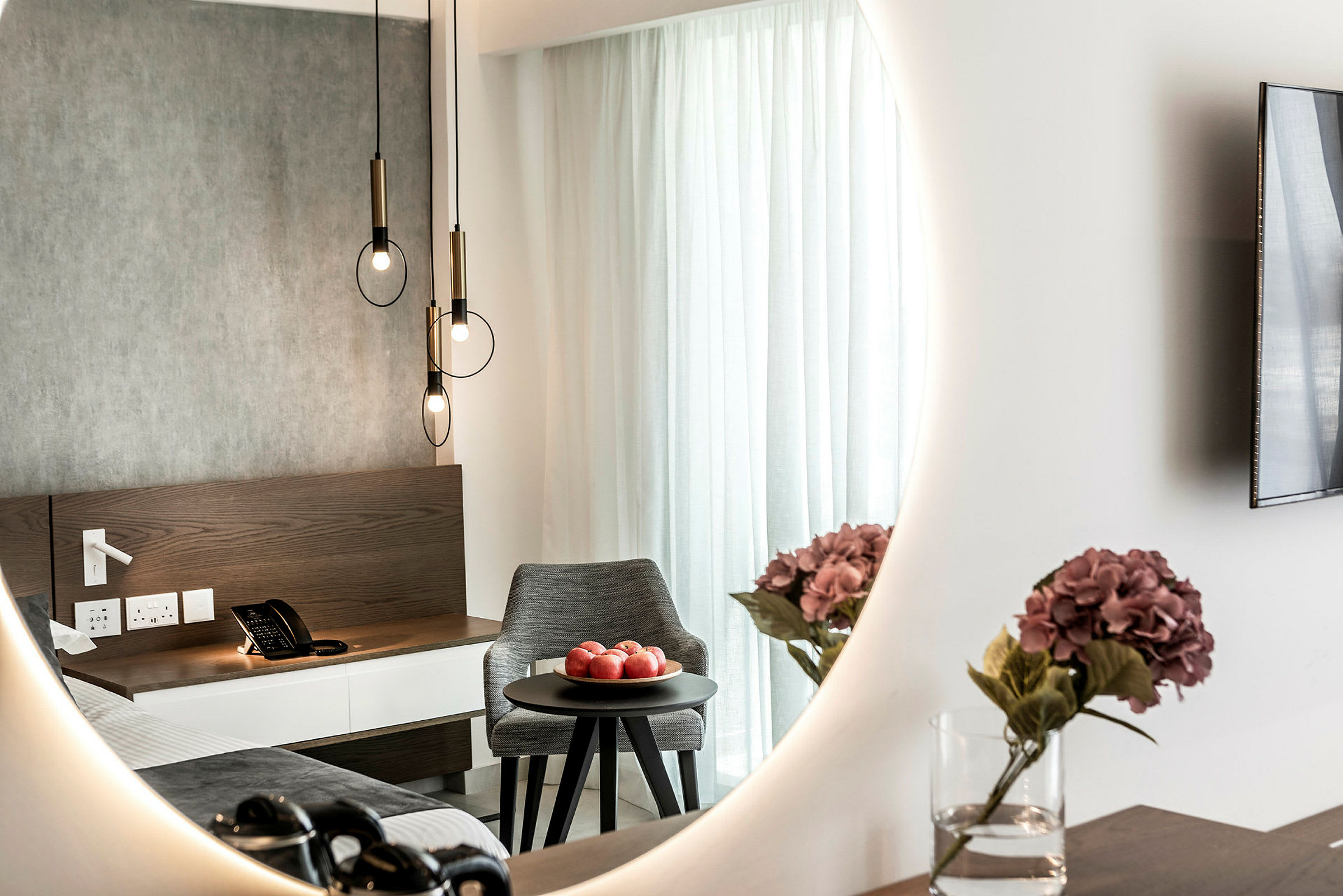 Hotellihuoneen sisustus, jossa peili heijastaa sänkyä, yöpöytää, jossa on puhelin, pyöreällä pöydällä olevaa kulhoa, jossa on punaisia omenoita, sekä maljakkoa, jossa on violetteja kukkia. (Tekoälyn luoma teksti)