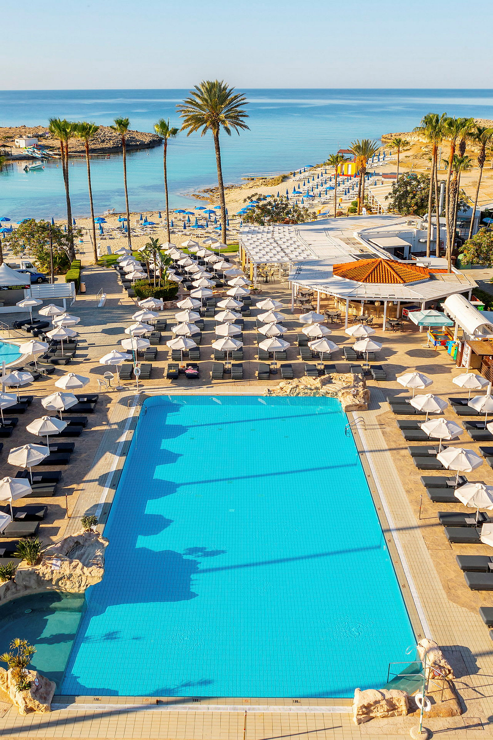 Kuva, jossa näkyy uima-allas aurinkotuoleilla ja aurinkovarjoilla ympäröitynä, sekä ranta ja meri taustalla. (Tekoälyn luoma teksti)