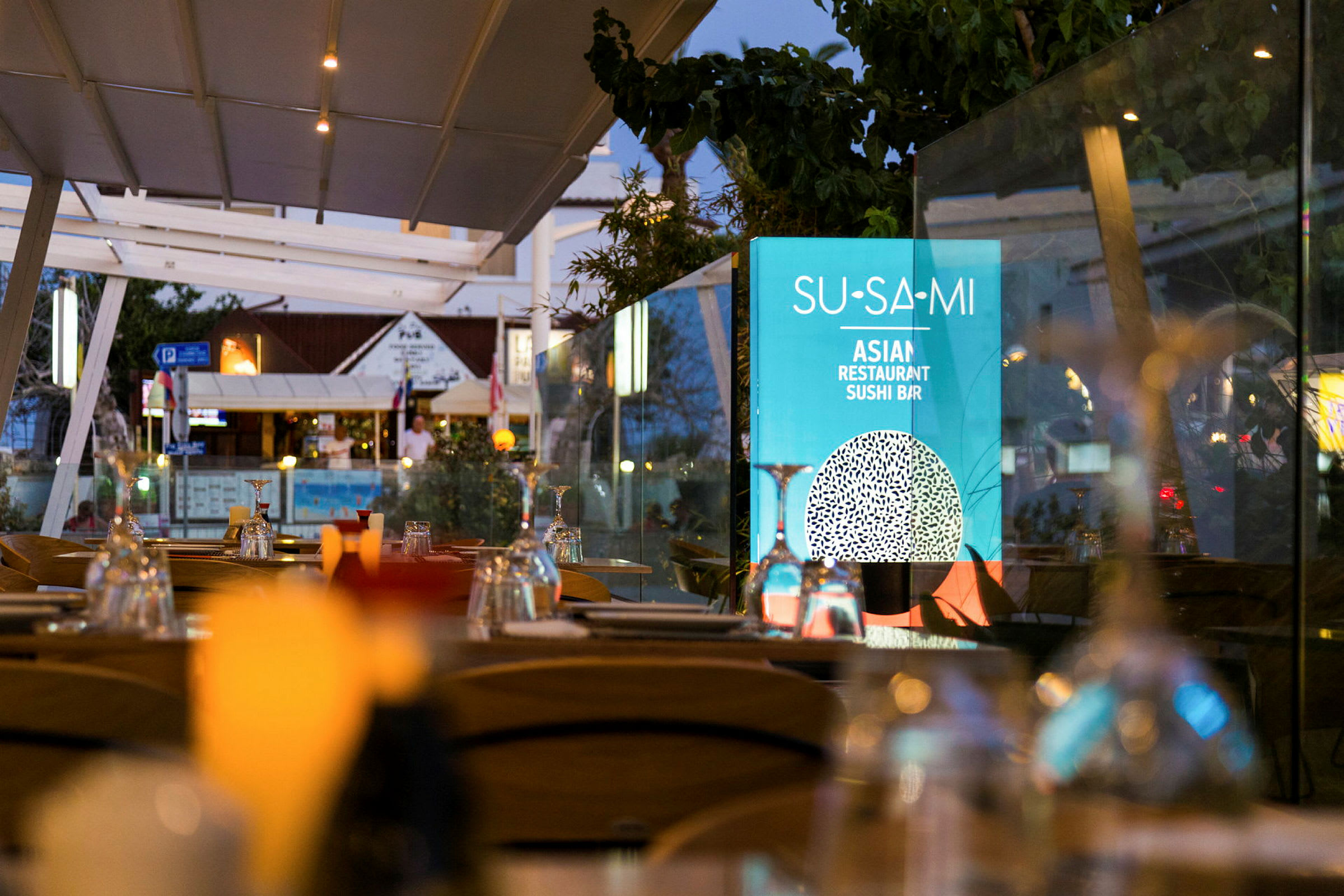Ulkoravintola, jossa on katettuja pöytiä ja kyltti, jossa lukee 'Su-Sa-Mi Asian Restaurant Sushi Bar'. (Tekoälyn luoma teksti)