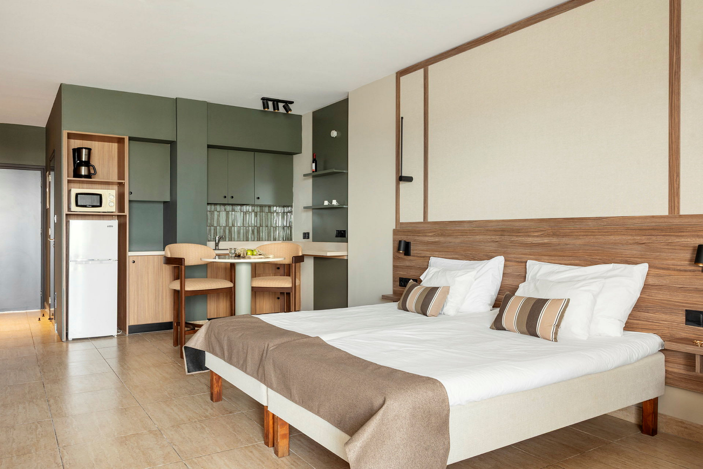 Sisäkuva valoisan ja modernin hotellihuoneen vuoteesta, pöydästä, tuoleista ja keittiötilasta. (Tekoälyn luoma teksti)