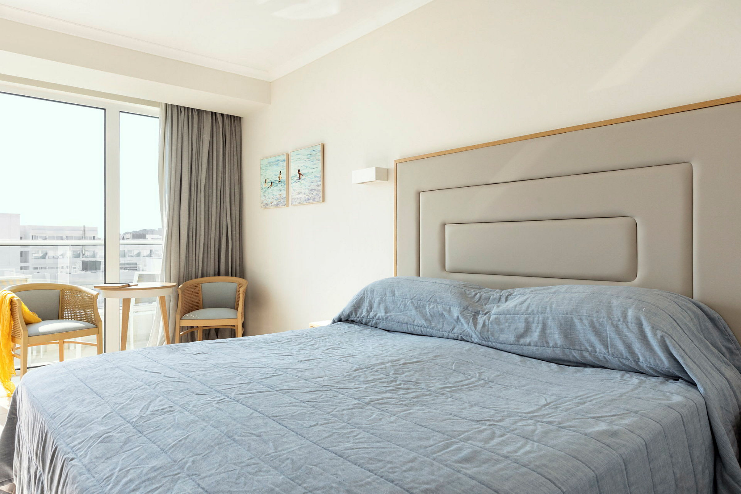 Hotellihuone, jossa sänky ja sininen päiväpeite, kaksi tuolia ja pöytä ikkunan vieressä. (Tekoälyn luoma teksti)