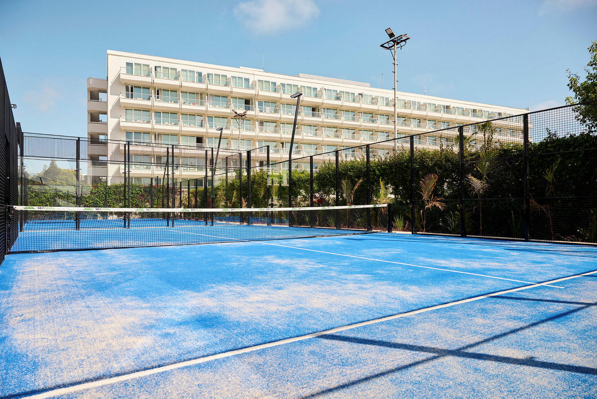 Padelkenttä, jossa sininen alusta, rakennuksen edessä, jossa on parvekkeita. (Tekoälyn luoma teksti)
