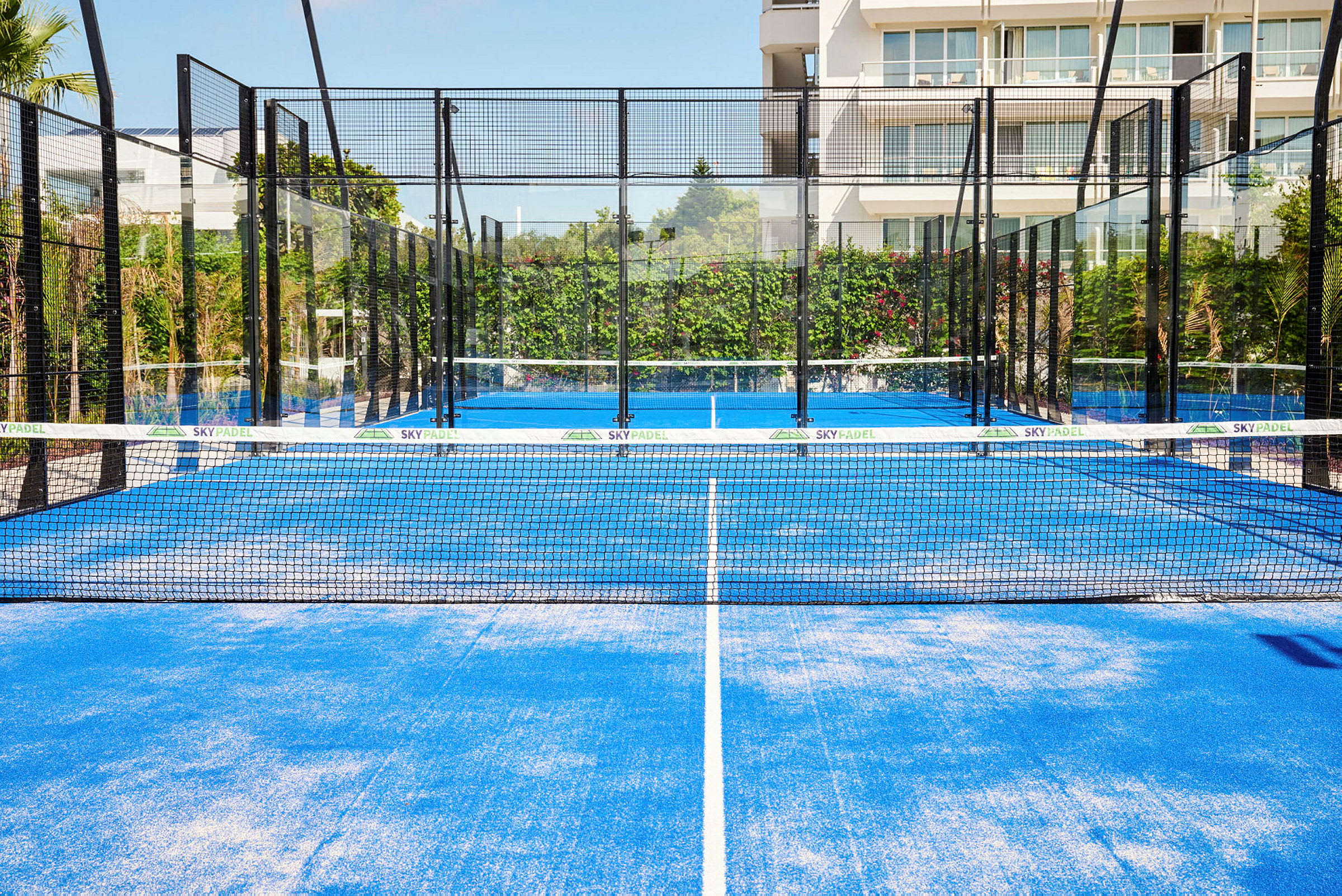 Sininen padelkenttä, jossa on verkko ja ympäröivät lasiseinät, sijaitsee ulkona kasvillisuuden ja rakennuksen taustalla. (Tekoälyn luoma teksti)