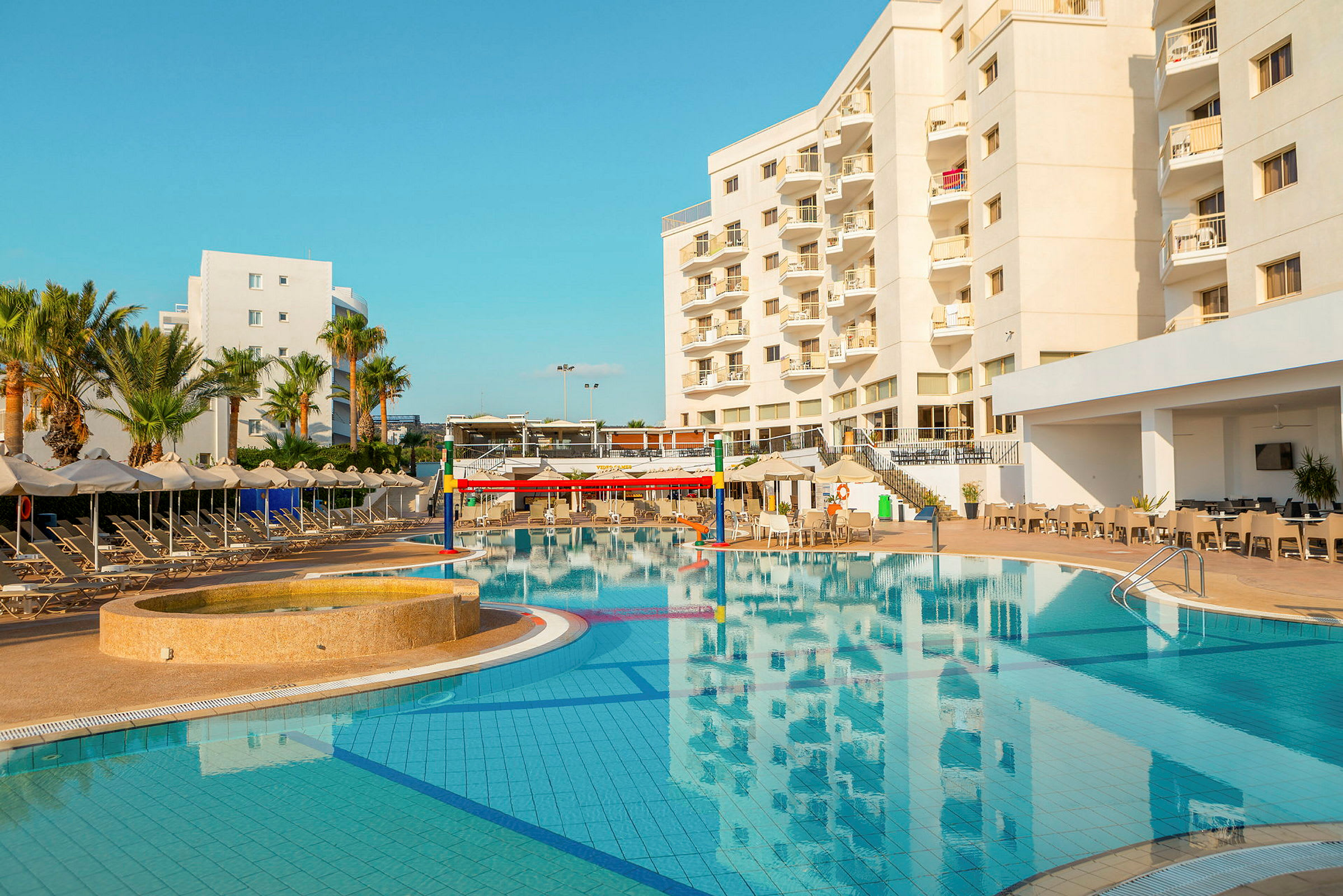 Hotellirakennus uima-altaan vieressä, jossa on aurinkotuoleja ja varjoja. (Tekoälyn luoma teksti)
