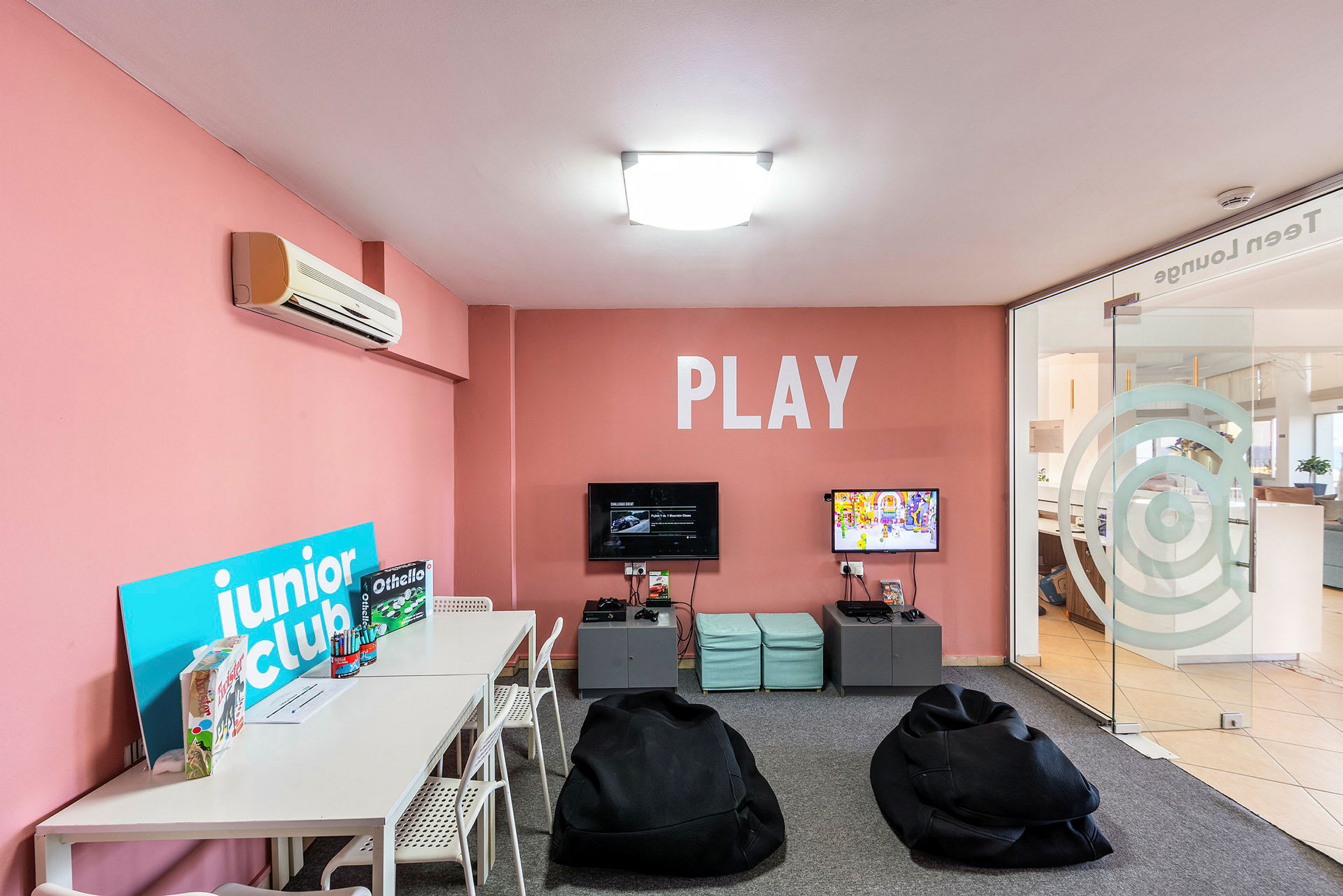 Leikkihuone, jossa on vaaleanpunaiset seinät, pöydät, tuolit, pelejä ja säkkituoleja sekä teksti 'PLAY' seinällä. (Tekoälyn luoma teksti)