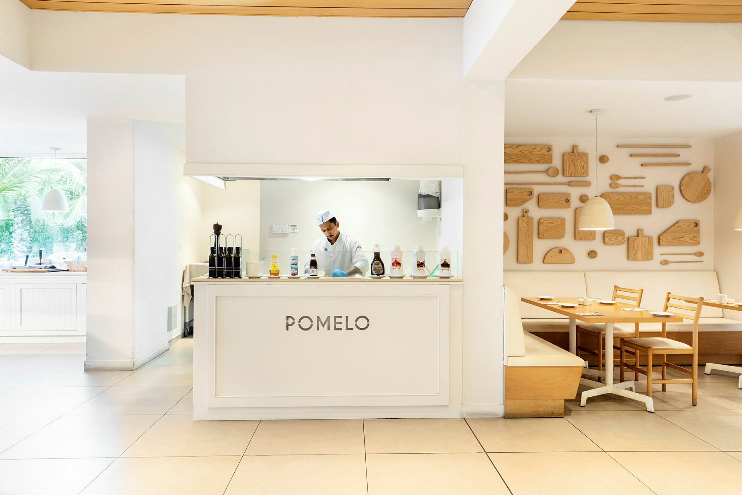 Ravintola nimeltään 'Pomelo', jonka keittiöalue on tiskin takana, sisustettu vaaleilla puupöydillä ja -tuoleilla sekä koristeellisilla puupaneeleilla seinällä. (Tekoälyn luoma teksti)