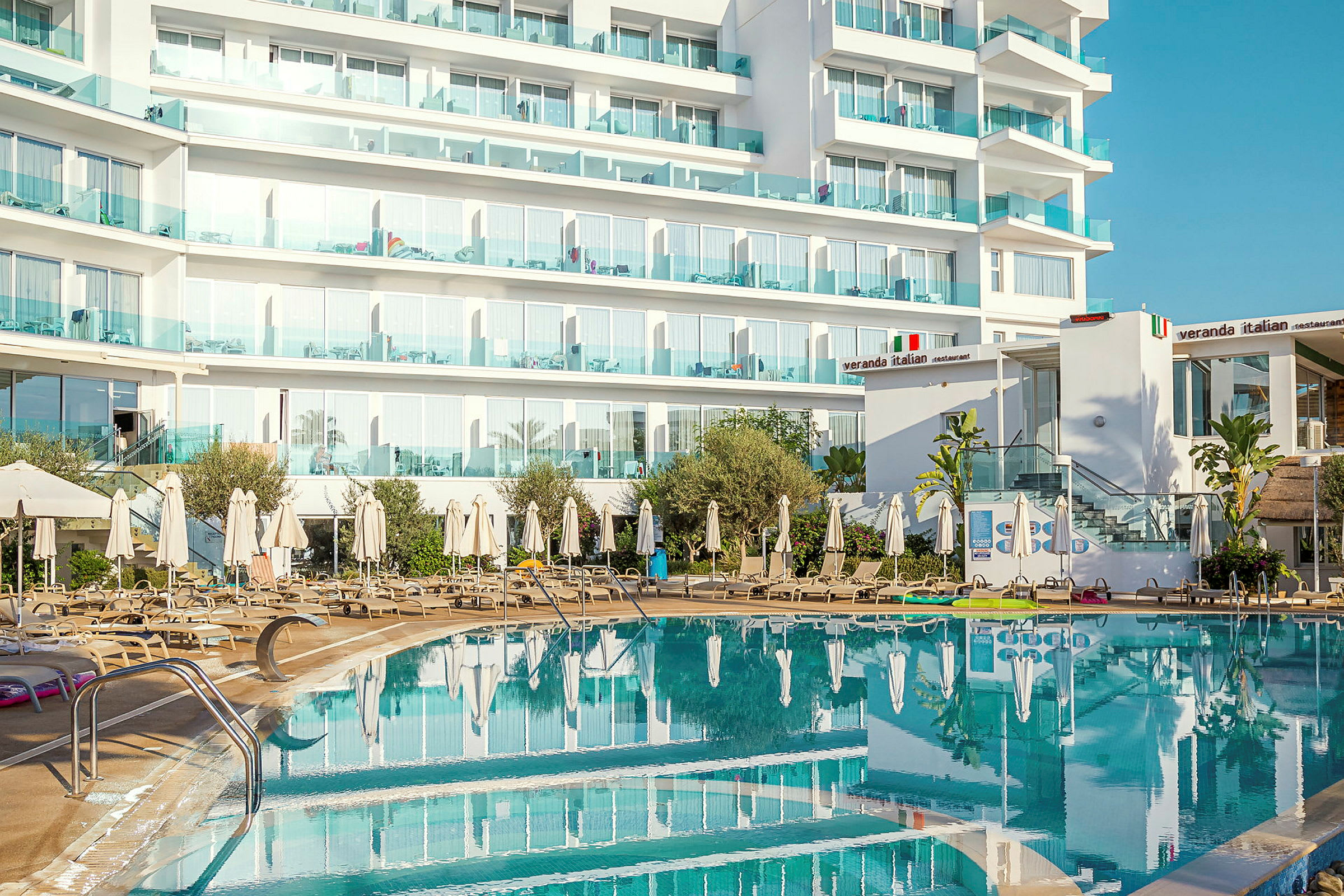 Hotellirakennus parvekkeineen ulkouima-altaan ja aurinkotuolien takana. (Tekoälyn luoma teksti)