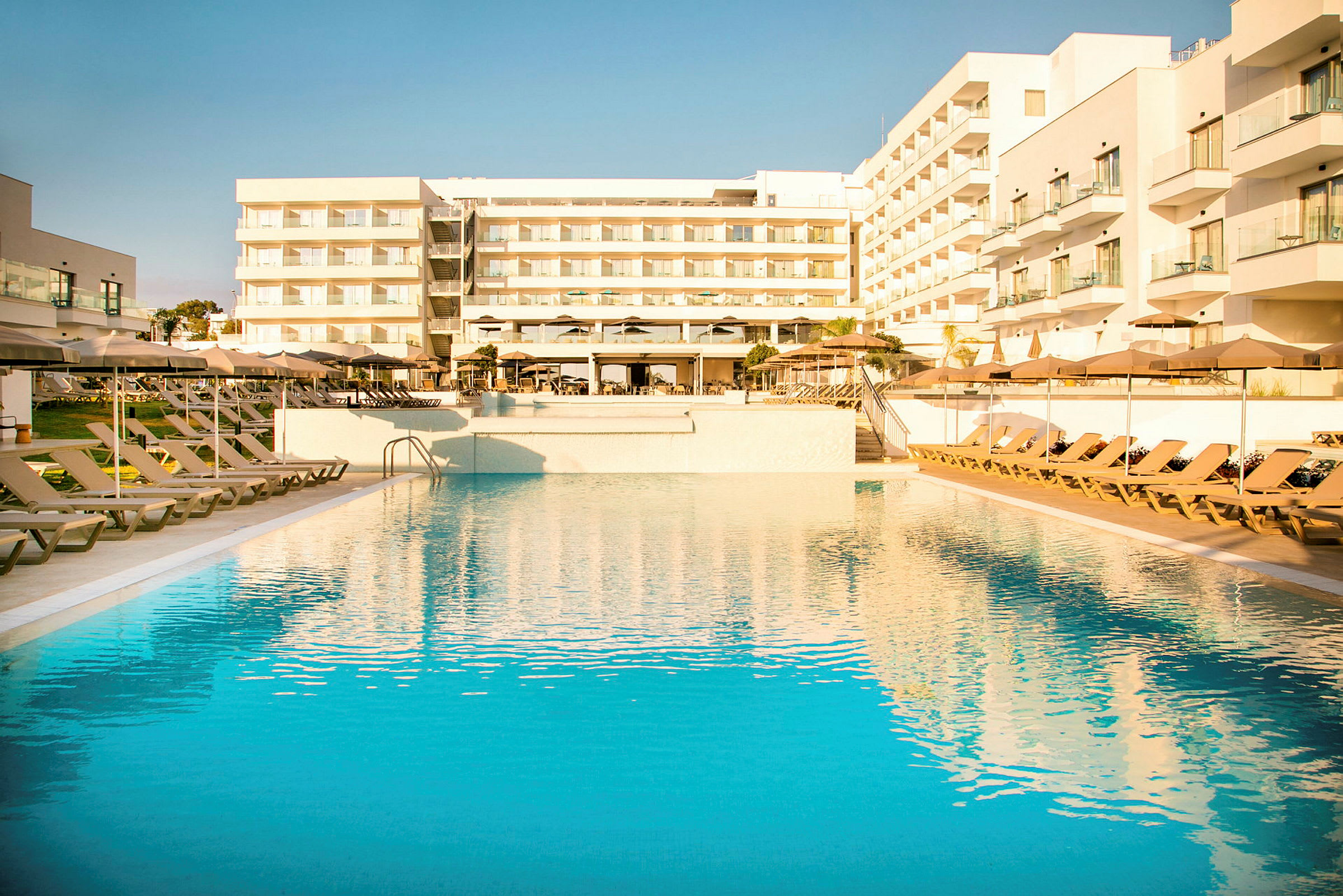 Suuri ulkouima-allas, jonka ympärillä on aurinkotuoleja ja aurinkovarjoja modernin hotellin edessä parvekkeineen. (Tekoälyn luoma teksti)