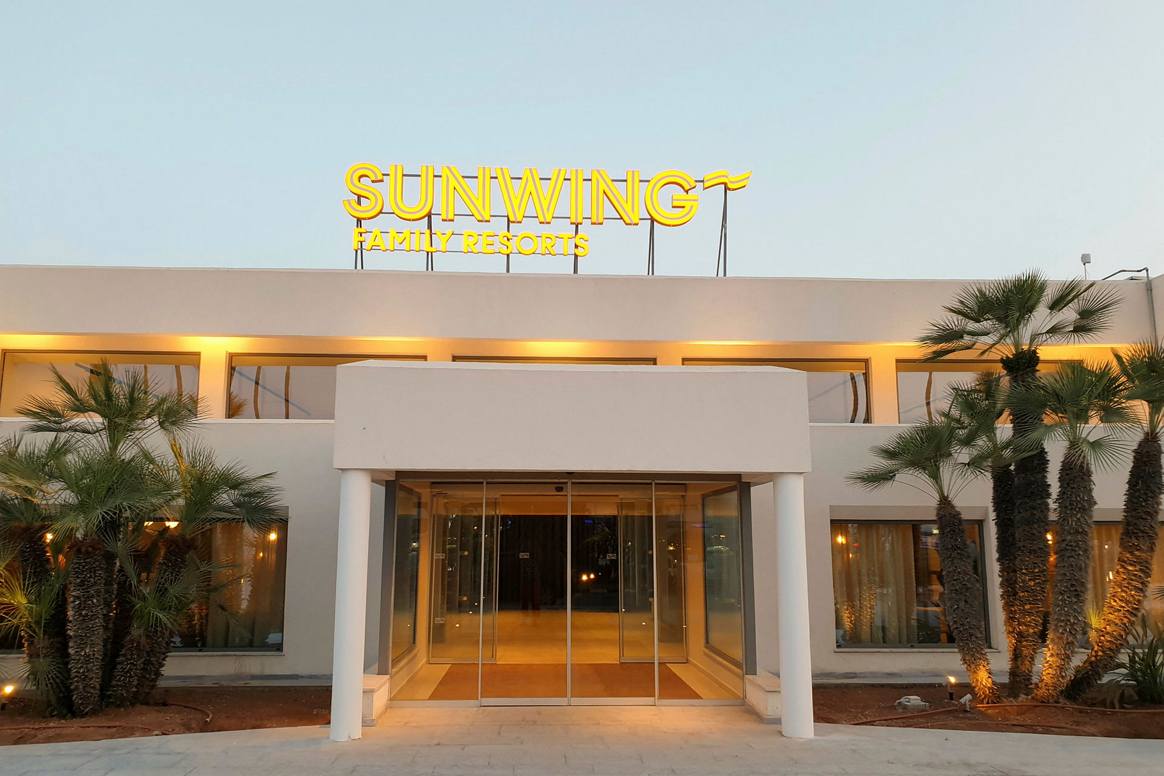 Rakennuksen etupuoli, jossa on taustavalaistu kyltti "Sunwing Family Resorts" ja ympärillä palmuja. (Tekoälyn luoma teksti)