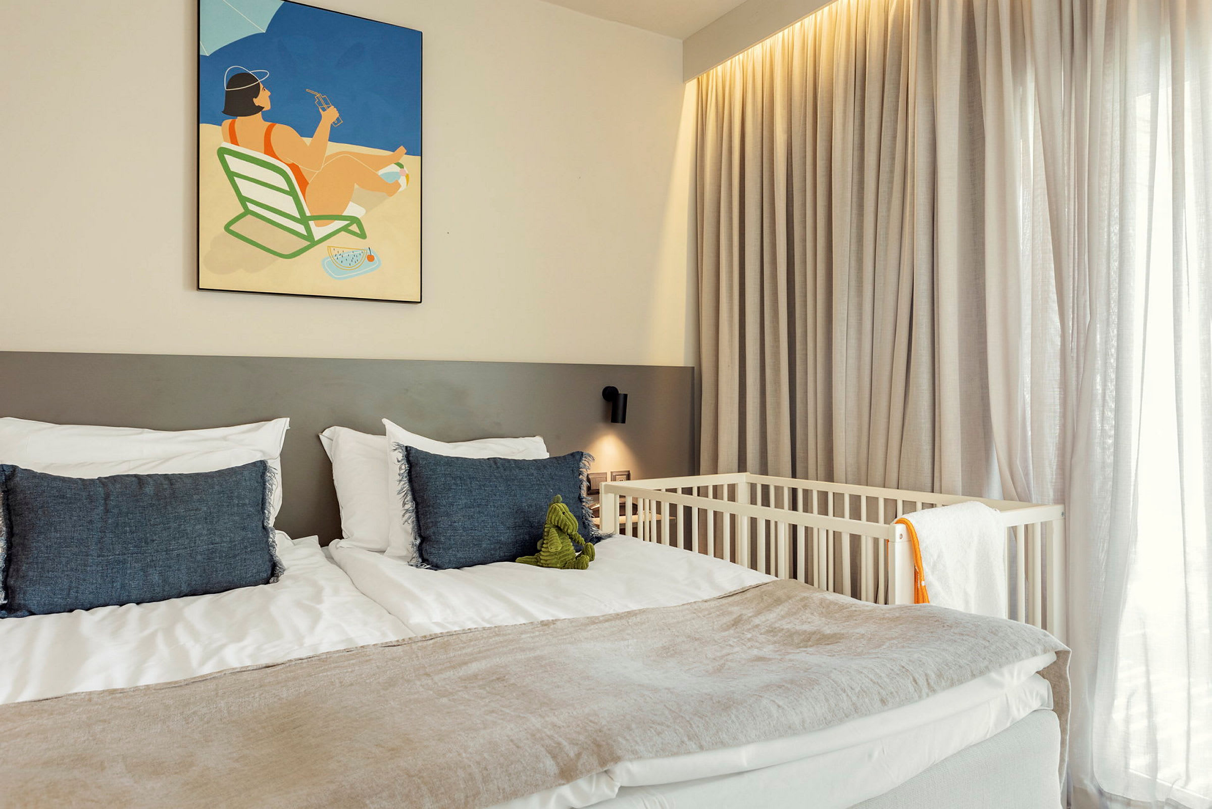 Hotellihuone, jossa on parisänky koristeltuna sinisillä tyynyillä ja seinällä yläpuolella oleva kuvitus. Vauvan pinnasänky näkyy ikkunan vieressä, jossa on ohuet verhot. (Tekoälyn luoma teksti)