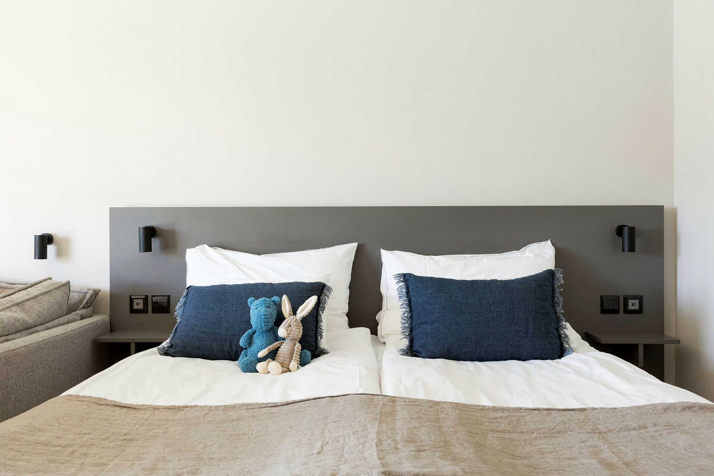 Kuvassa on moderni makuuhuone, jossa on parivuode valkoisilla vuodevaatteilla ja sinisillä tyynyillä. Sängyllä on kaksi pehmolelua, yksi sininen ja yksi beige. Seinälamput on kiinnitetty sängyn päätyyn. (Tekoälyn luoma teksti)