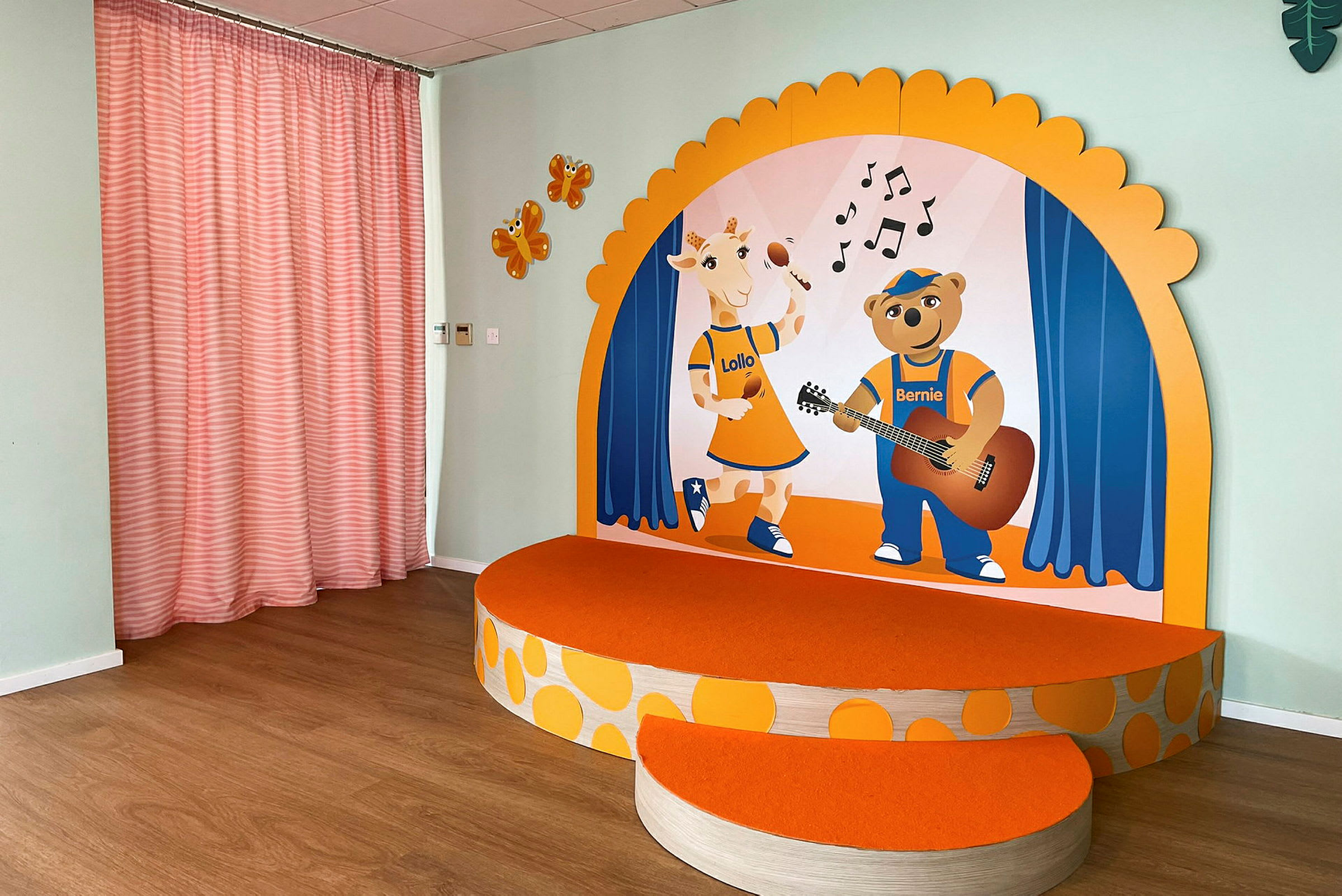 Pieni korotettu lava, jossa on oranssi pinta ja piirroksia kirahvista ja karhusta musiikkiteemaisella taustalla. Sivuseinällä on oransseja kuvioita ja vaaleanpunaiset verhot. (Tekoälyn luoma teksti)