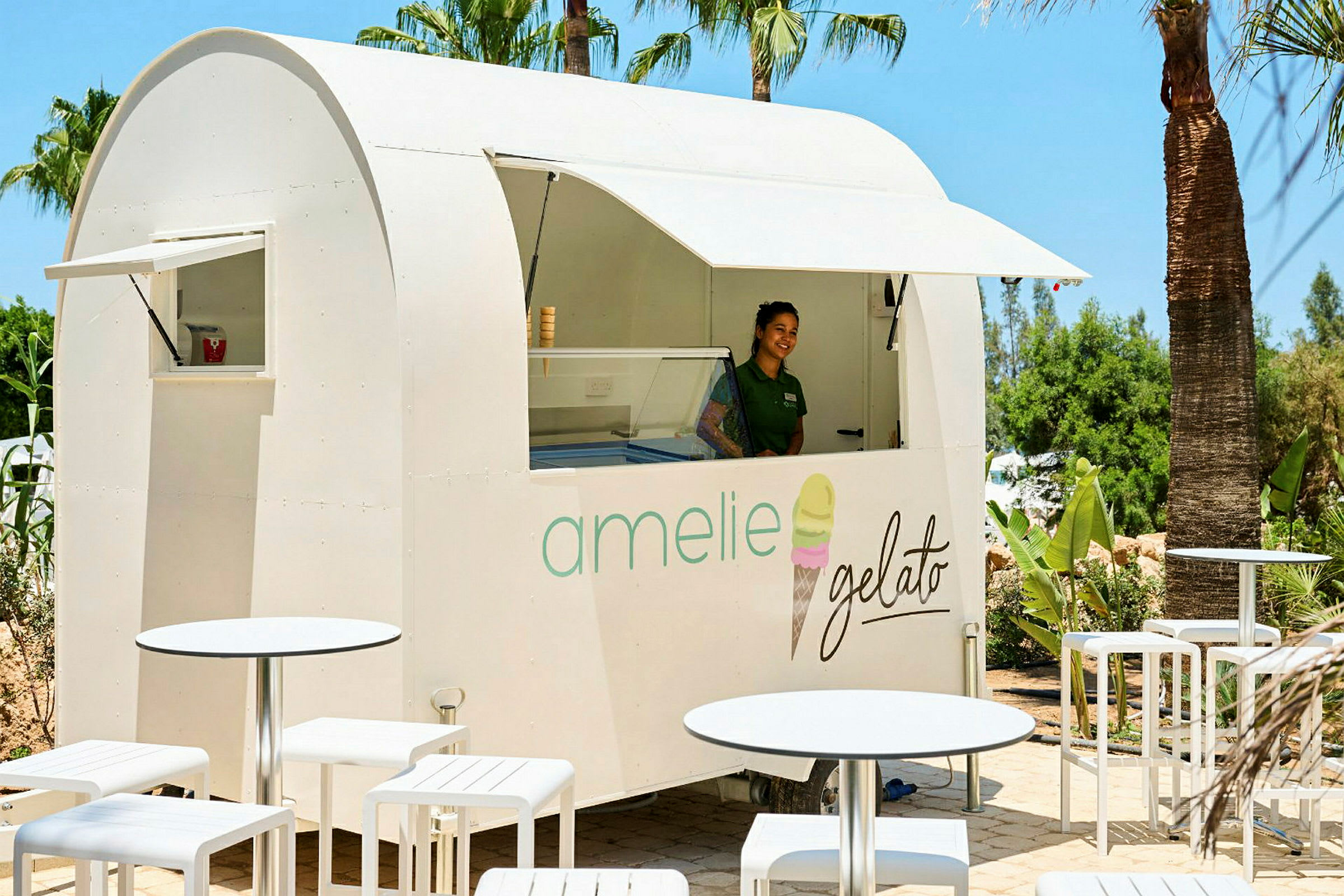 Valkoinen jäätelökioski, jossa lukee "amelie gelato", seisoo palmujen keskellä, ympärillä valkoisia ulkokalusteita. (Tekoälyn luoma teksti)