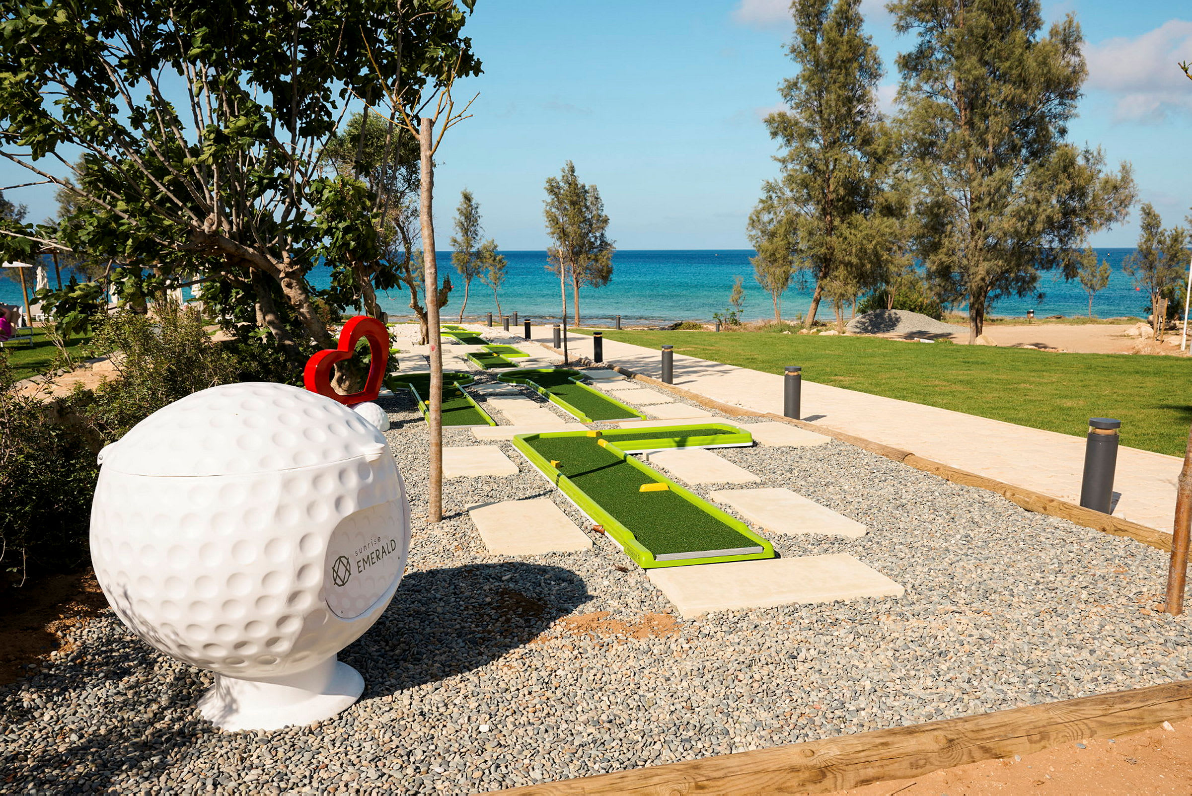 Minigolf-rata, jossa on vihreitä ratoja, sijaitsee lähellä rantaa sinisen meren taustalla. Suuri golfpalloveistos näkyy vieressä. (Tekoälyn luoma teksti)