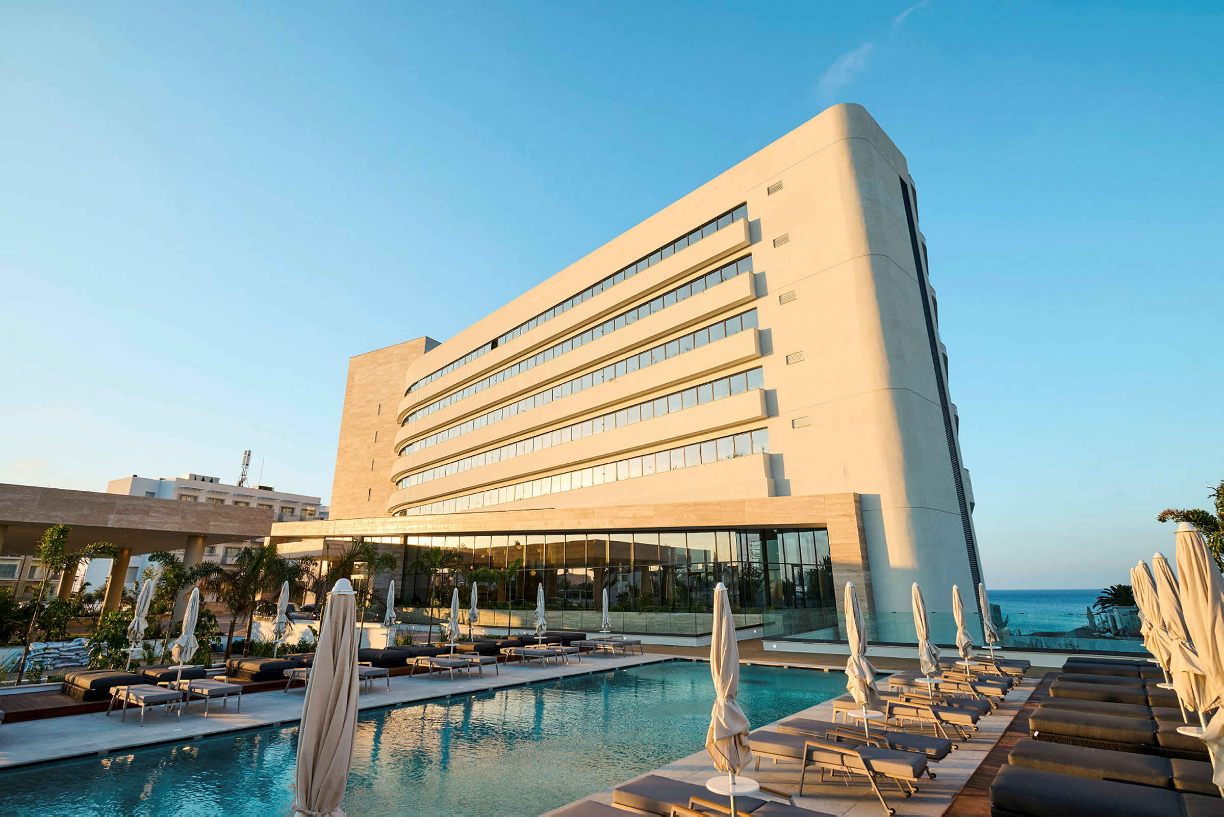 Moderni hotellirakennus uima-altaan vieressä, jossa on aurinkotuoleja ja aurinkovarjoja. Meri näkyy taustalla. (Tekoälyn luoma teksti)