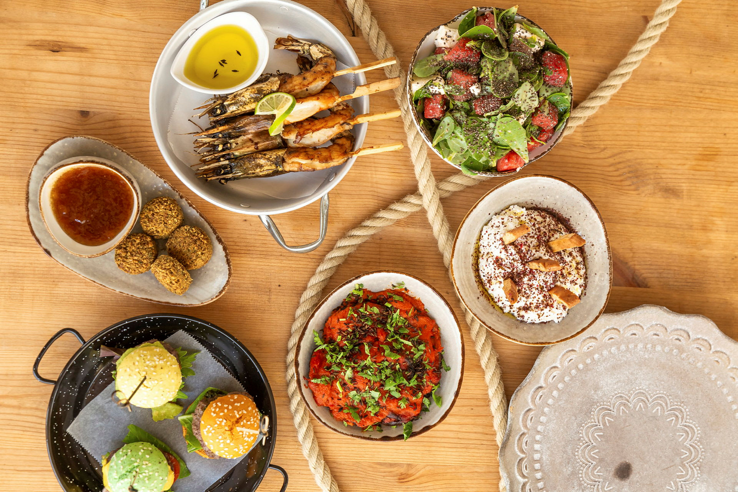 Monenlaisia ruokalajeja, kuten vartaan päällä olevat katkaravut, salaatti, falafel, hampurilaisia, sekä dippejä ja lisukkeita puupöydällä. (Tekoälyn luoma teksti)
