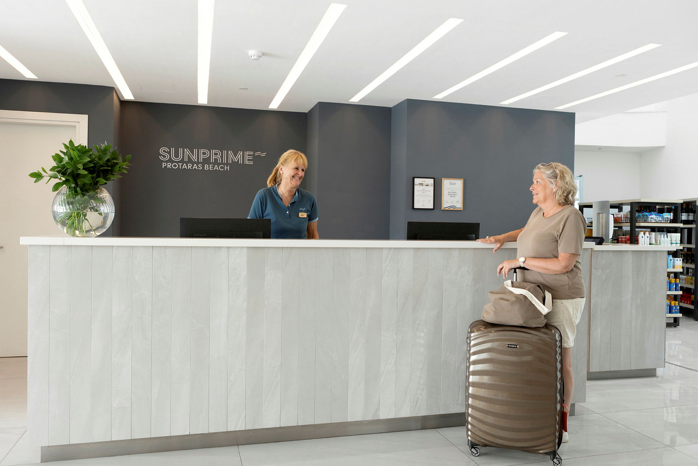 Sunprime Protaras Beach -hotellin vastaanotto modernilla sisustuksella, sisältäen tiskin, jonka edessä matkalaukku ja käsilaukku. (Tekoälyn luoma teksti)
