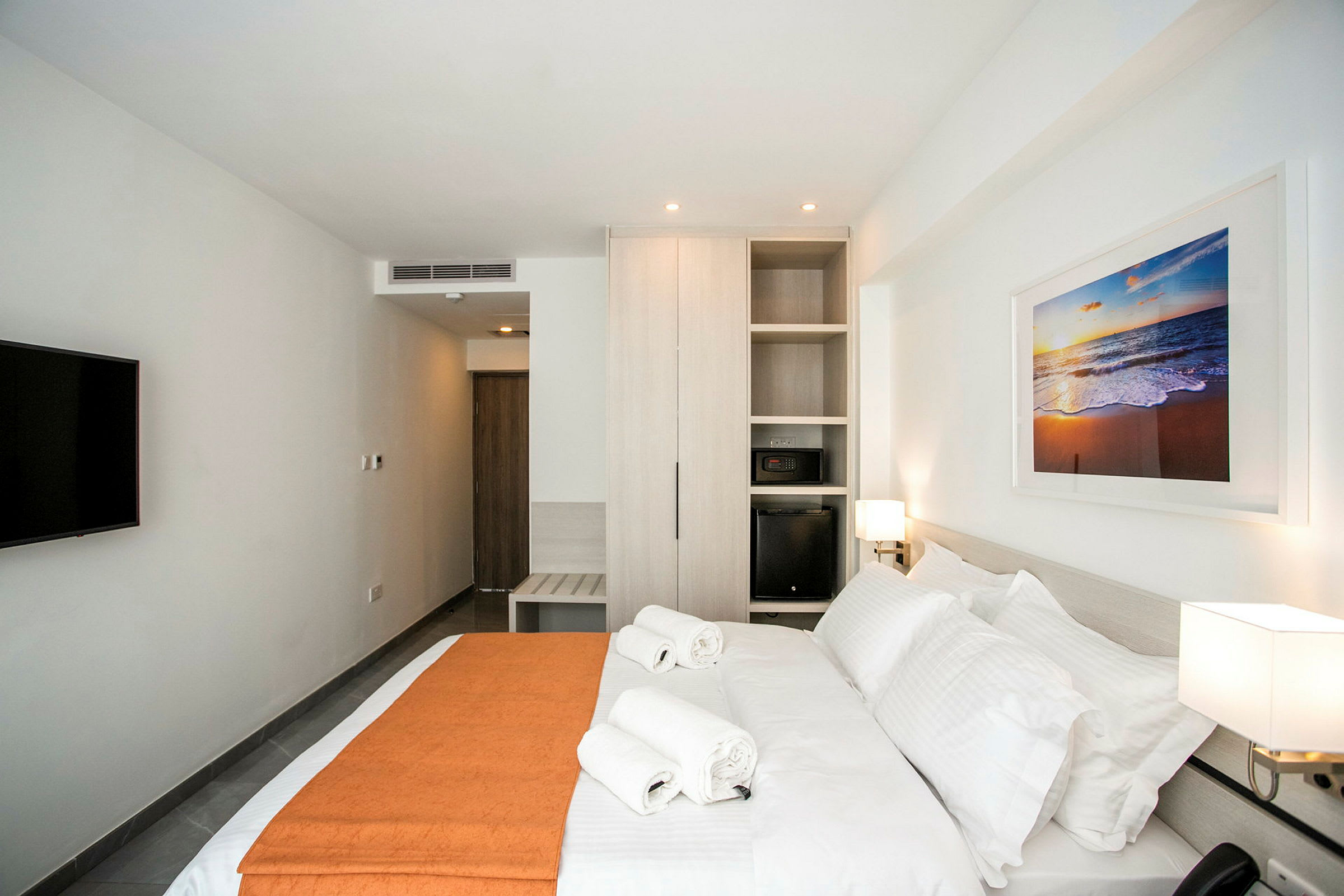 Moderni hotellihuone, jossa on suuri sänky, kaksi pyyhettä sen päällä, vaatekaappi, seinälle kiinnitetty televisio ja taulu, jossa on rantamaisema. (Tekoälyn luoma teksti)