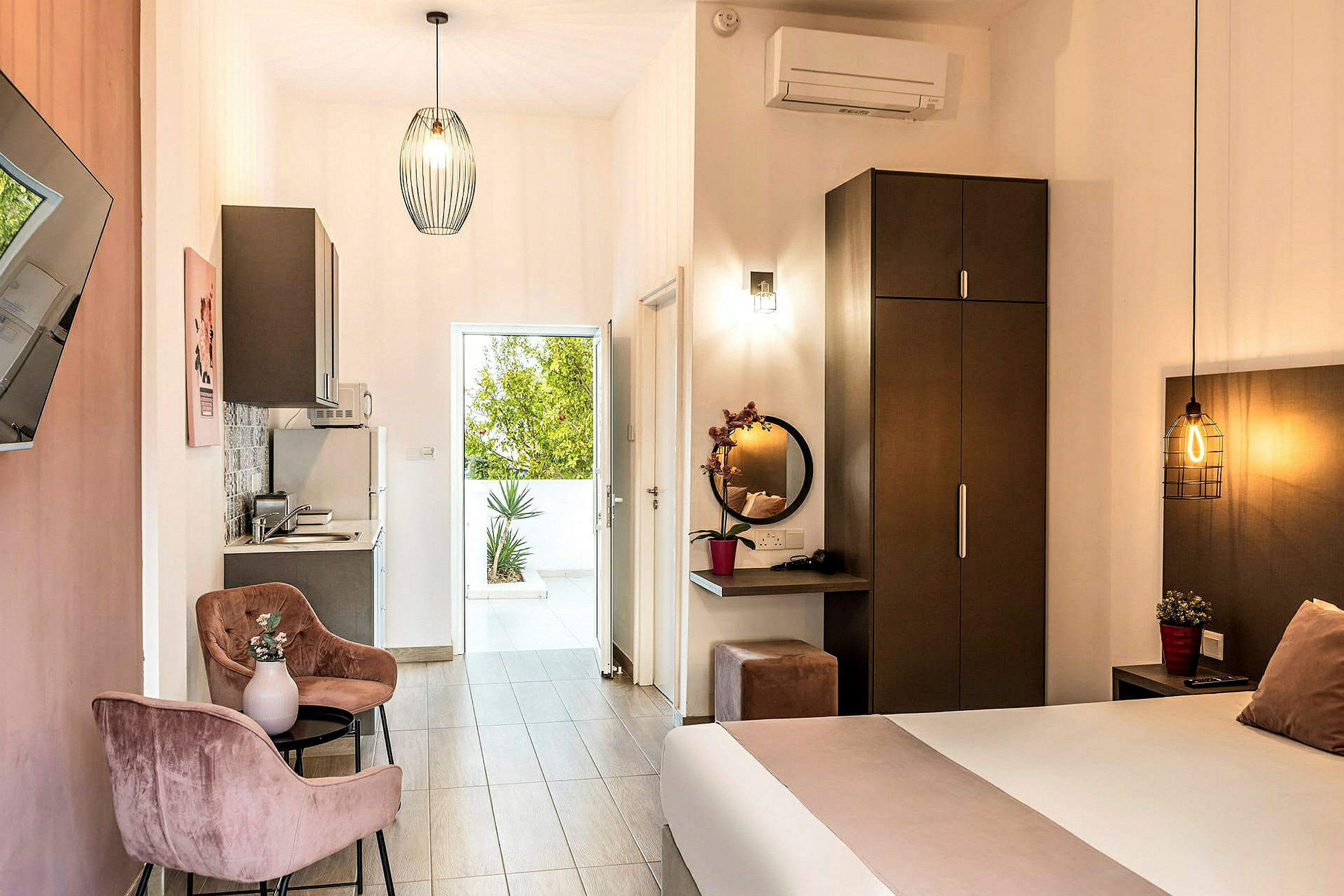 Hotellihuone jossa on sänky, työpöytä peilillä, vaatekaappi ja pieni keittiö. Ovi johtaa terassille, jossa on vehreyttä. (Tekoälyn luoma teksti)