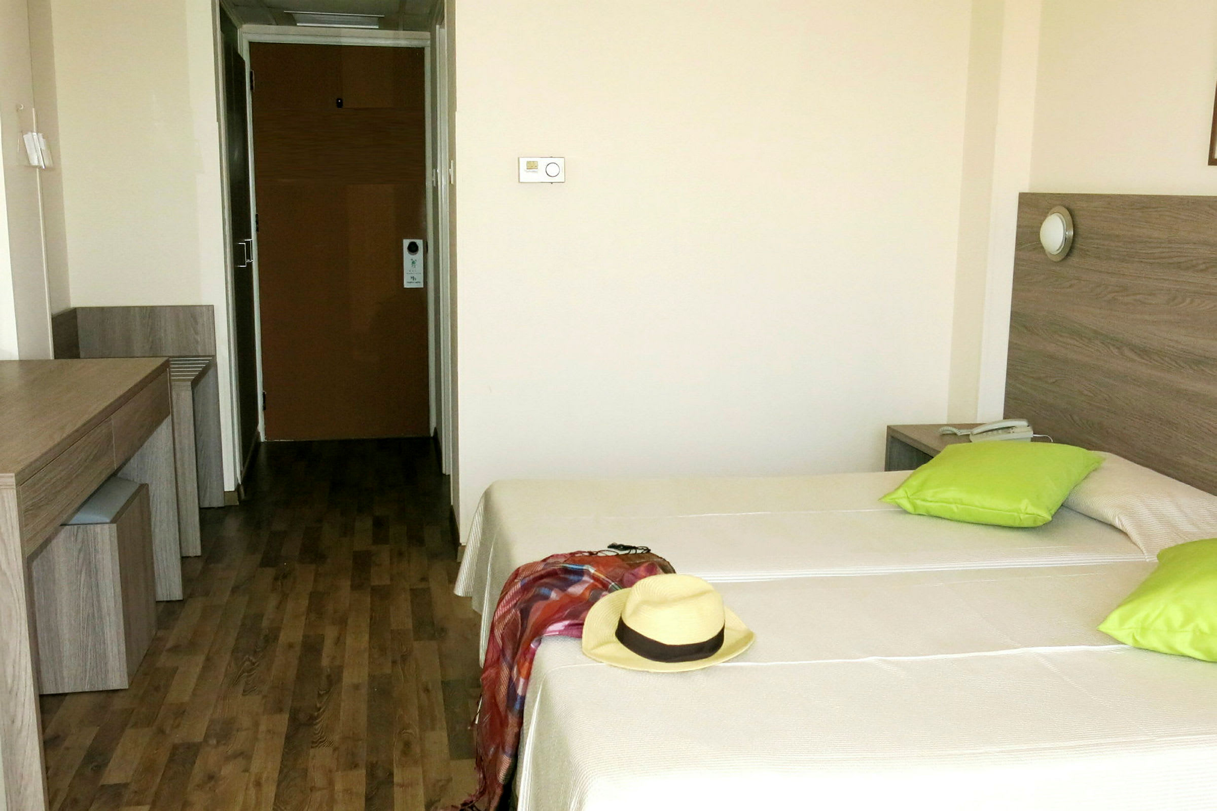 Hotellihuone, jossa kaksi yhden hengen vuodetta, vihreät tyynyt sekä hattu ja huivi toisella vuoteella. (Tekoälyn luoma teksti)