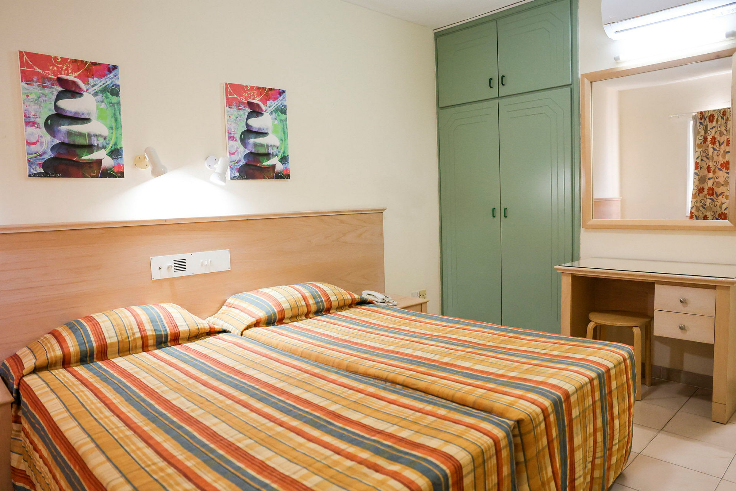 Hotellihuone, jossa on kaksi yksittäistä vuodetta ja ruudulliset päiväpeitteet. Siellä on vihreä vaatekaappi, peili työpöydän yläpuolella ja taidemaalauksia seinällä. (Tekoälyn luoma teksti)