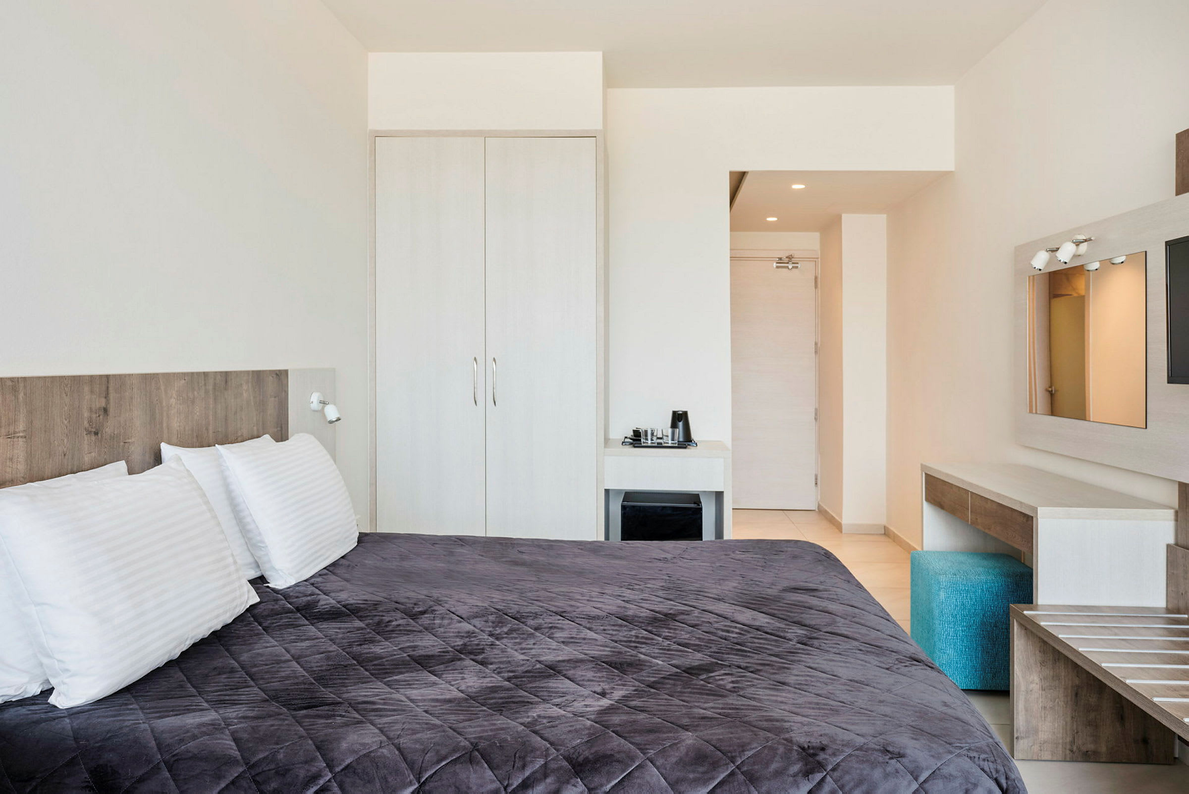 Hotellihuone, jossa on suuri sänky, tumma harmaa päiväpeite ja valkoiset tyynyt. Näkyy myös kaappi, työpöytä kahvikoneella sekä tuoli. (Tekoälyn luoma teksti)