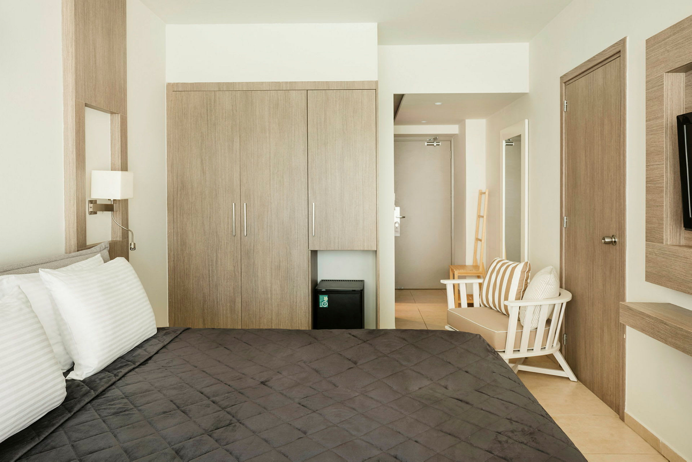 Hotellihuone, jossa on sänky, vaatekaappi, tuoli ja pieni jääkaappi vaatekaapin alla. (Tekoälyn luoma teksti)