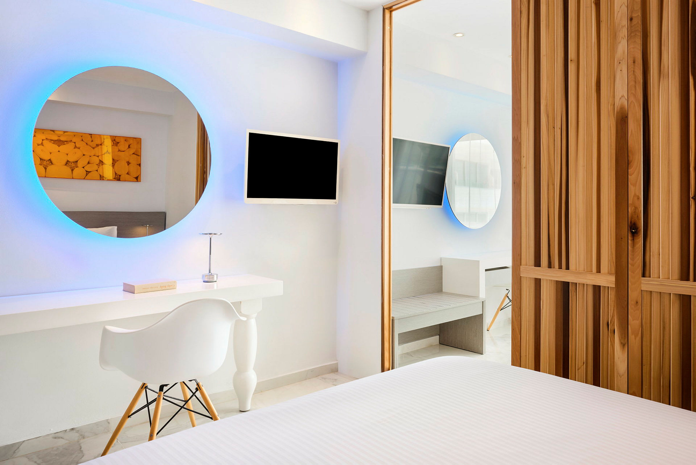 Moderni hotellihuone, jossa on pyöreä valaistu peili, TV-näyttö, työpöytä ja tuoli. Seinät ovat valkoiset ja huonekaluissa on minimalistinen tyyli. (Tekoälyn luoma teksti)