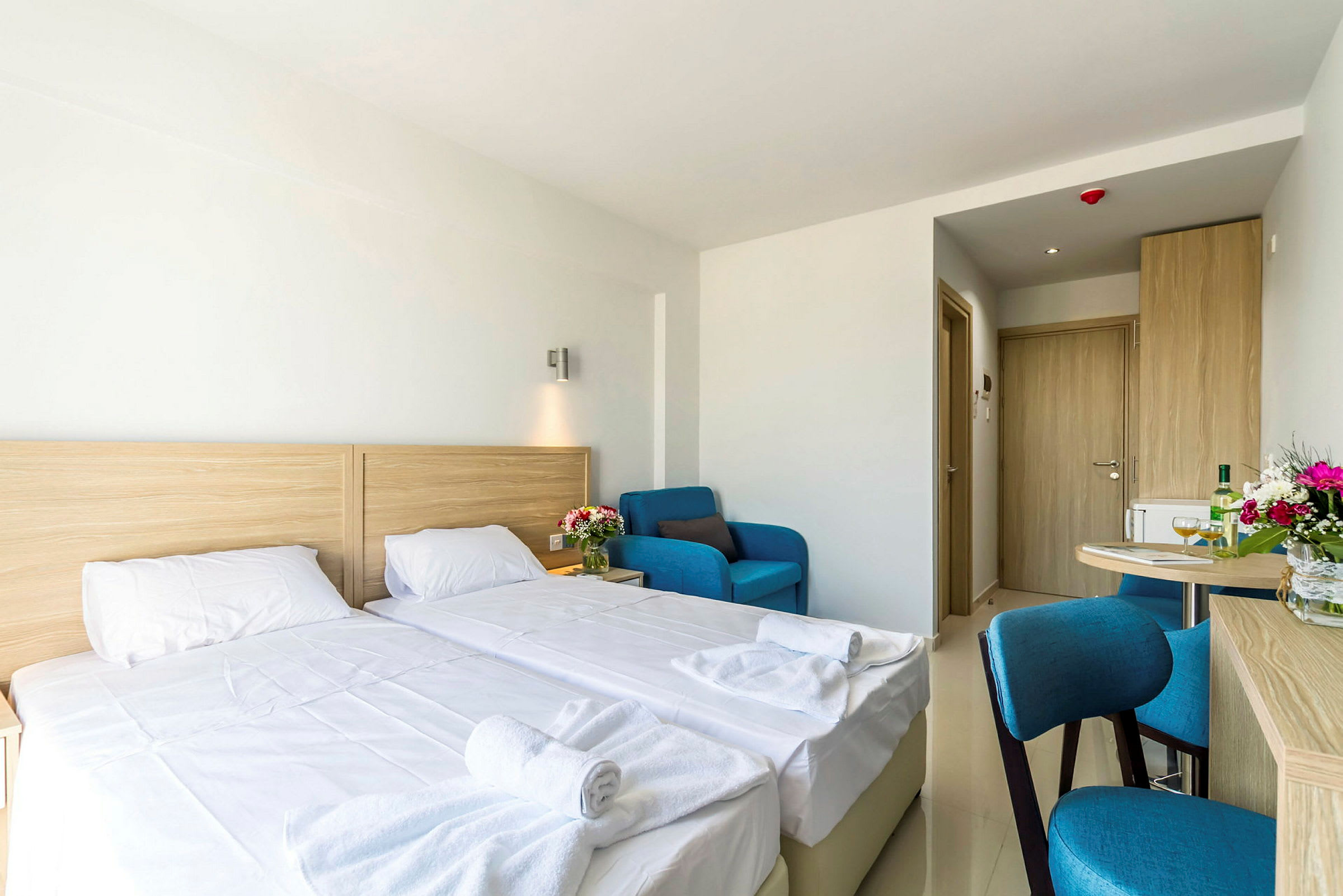 Hotellihuone, jossa on kaksi erillistä sänkyä, sininen nojatuoli, pöytä ja ovi taustalla. (Tekoälyn luoma teksti)