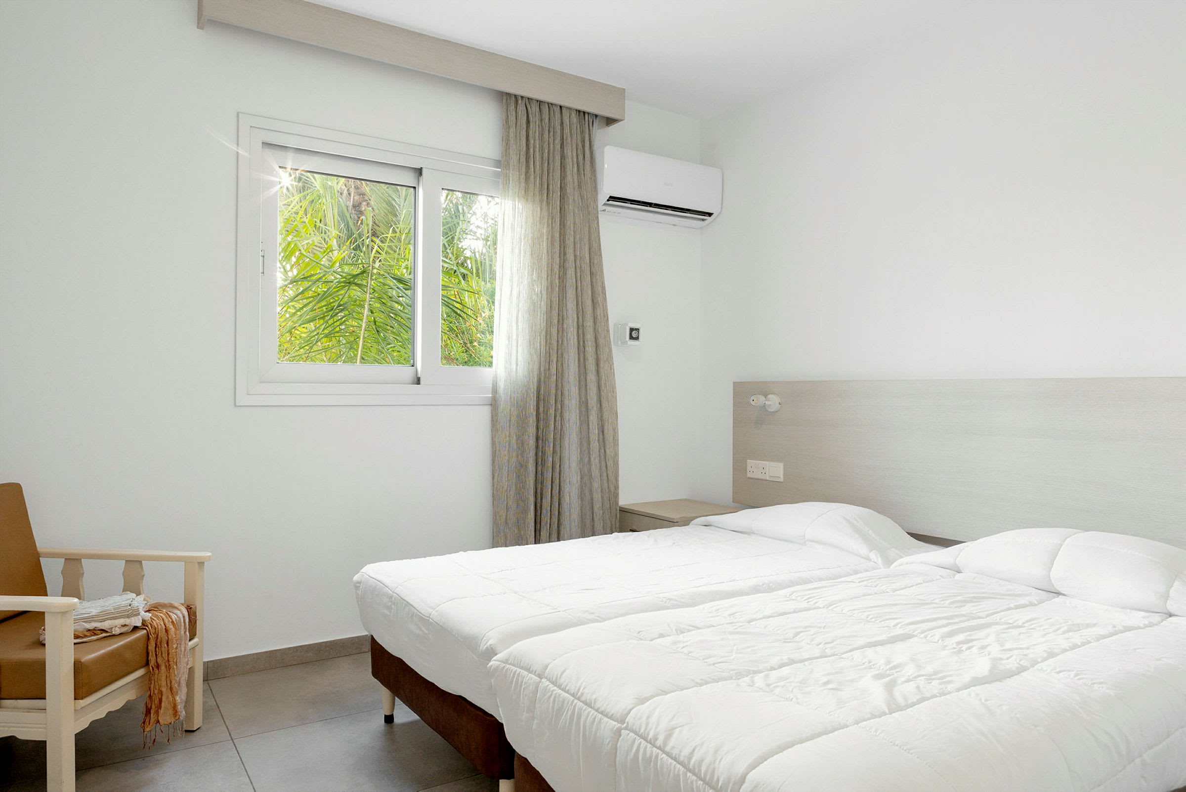 Hotellihuone, jossa kaksi erillistä vuodetta, nojatuoli ja ikkuna näkymällä vehreyteen. (Tekoälyn luoma teksti)