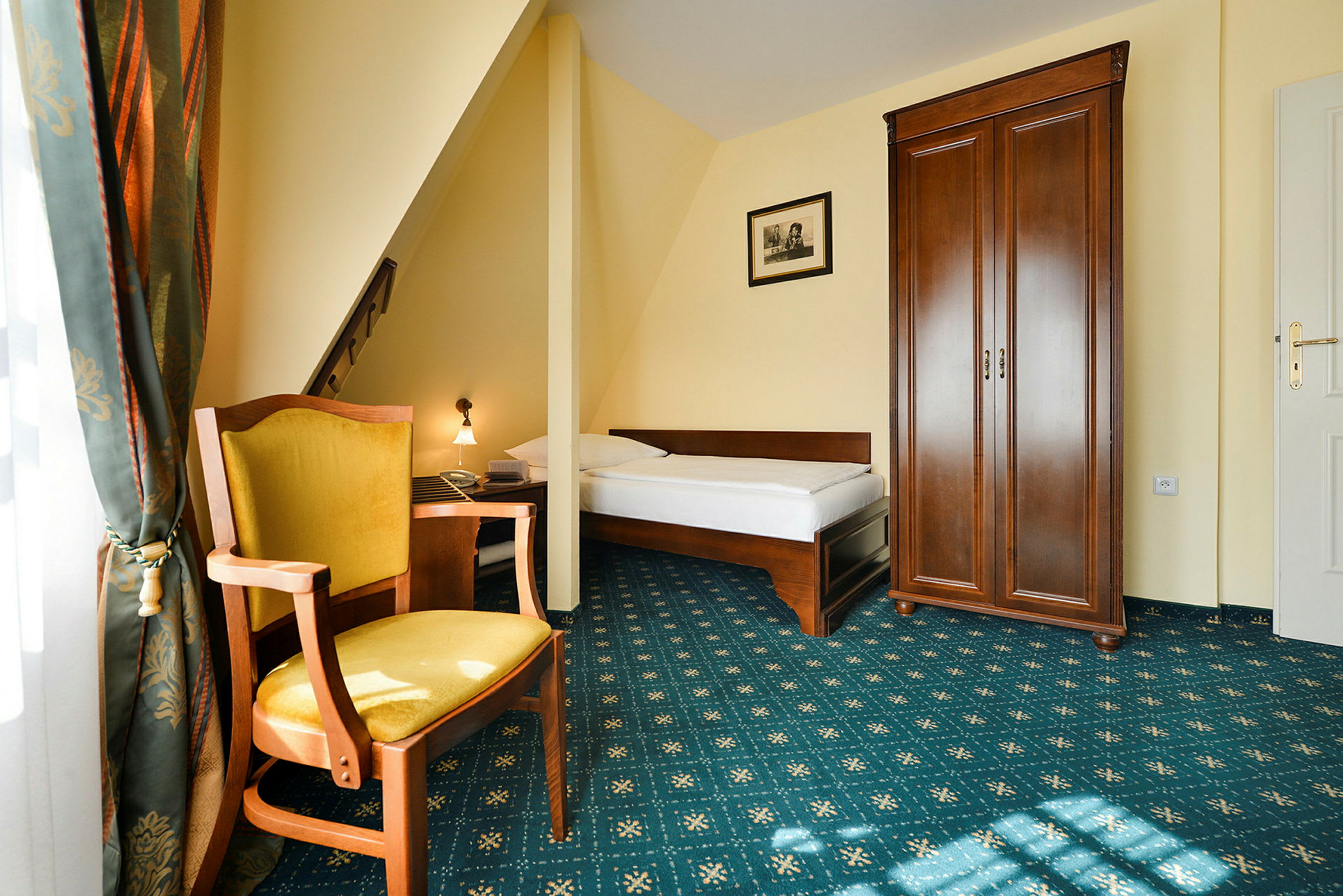 Hotellihuone, jossa on yhden hengen sänky, puinen tuoli, jossa on keltainen istuin, korkea vaatekaappi ja ikkuna verhoineen. (Tekoälyn luoma teksti)