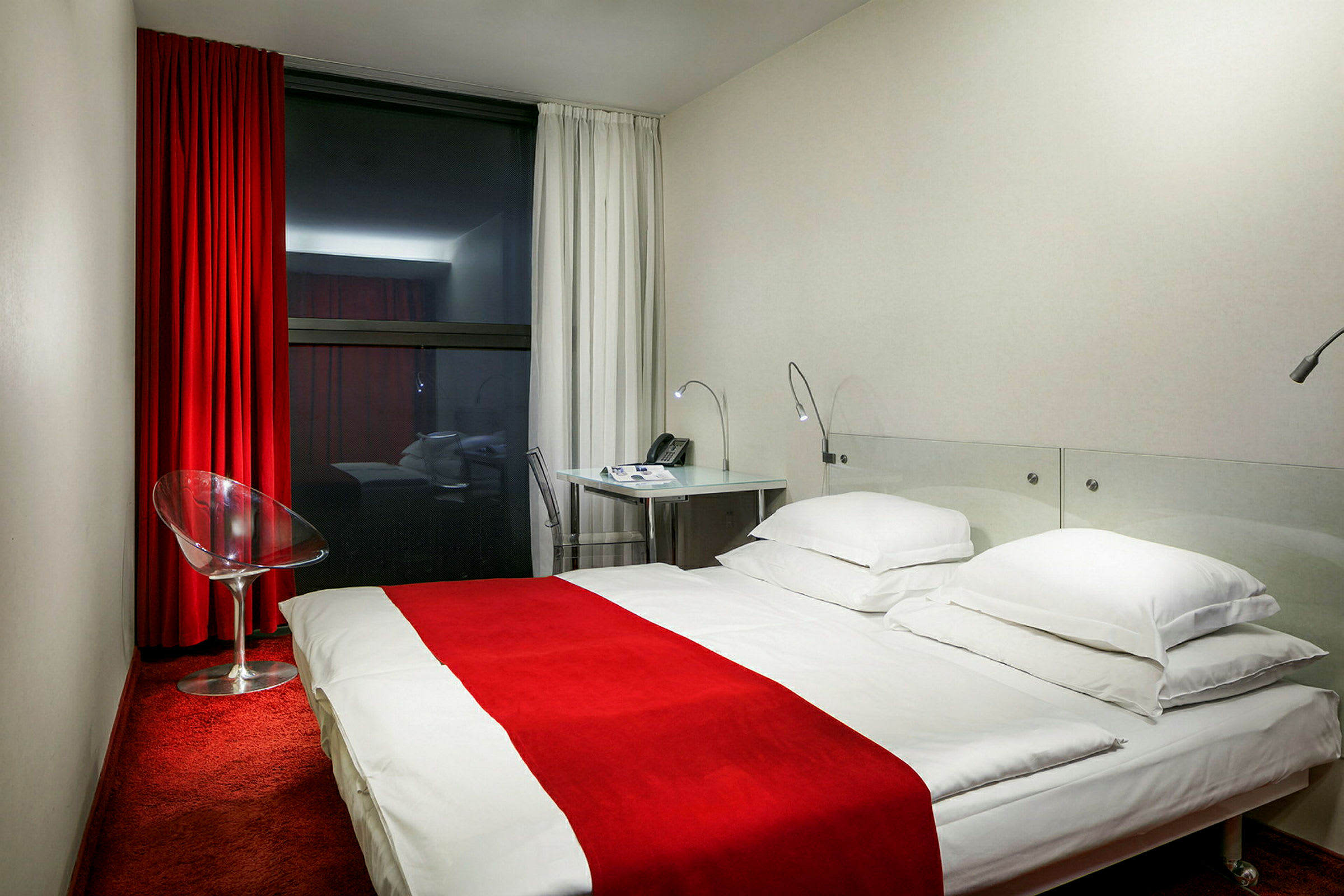 Hotellihuone, jossa on punainen matto, valkoinen sänky punaisella päiväpeitteellä, lasituoli ja kirjoituspöytä. (Tekoälyn luoma teksti)