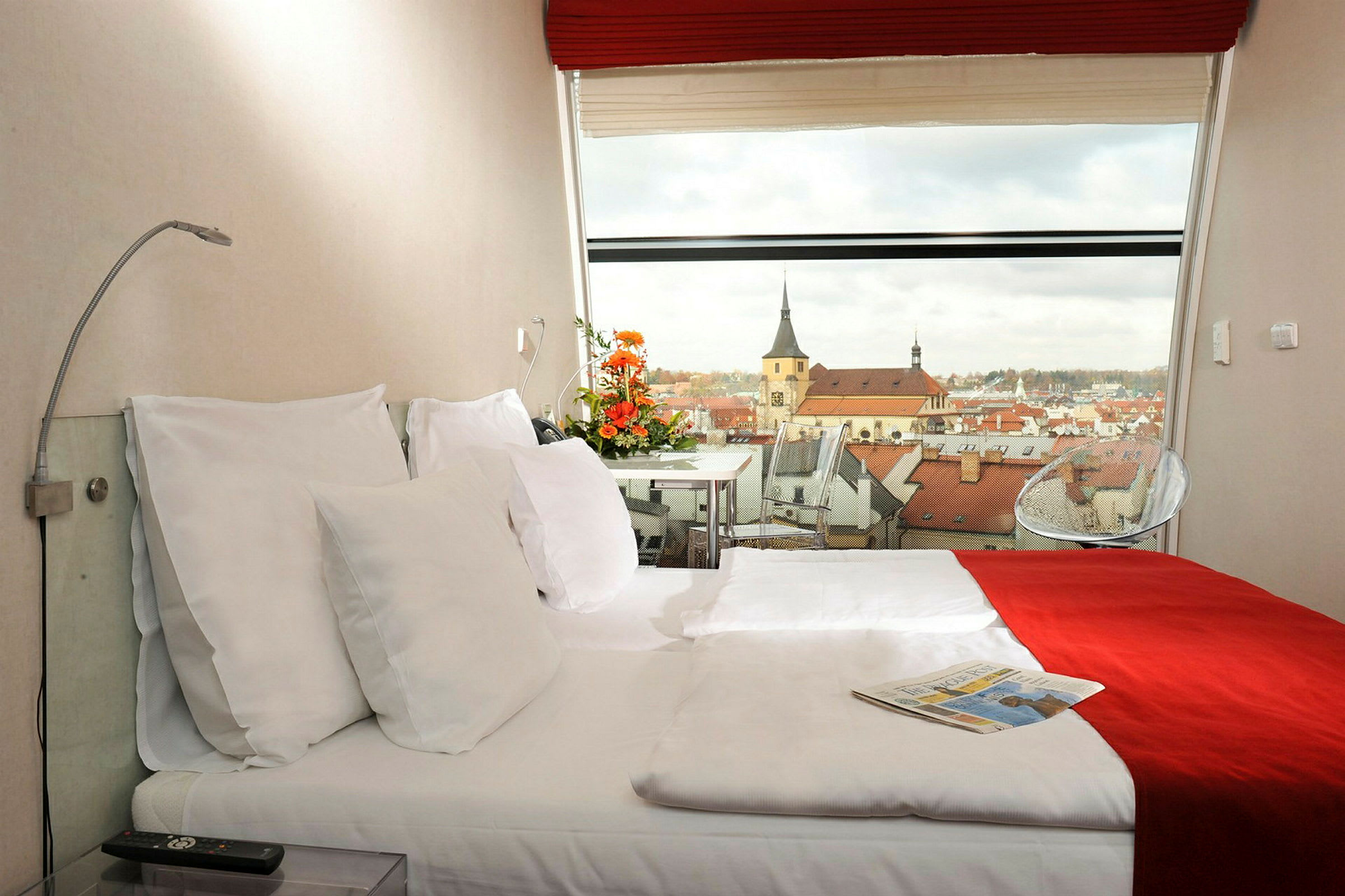 Hotellihuone, jossa on parivuode, koristeltu punaisella ja valkoisella vuodevaatteella. Panoraamanäkymä kaupunkiin, jossa on kirkon rakennuksia, näkyy ikkunan läpi. (Tekoälyn luoma teksti)