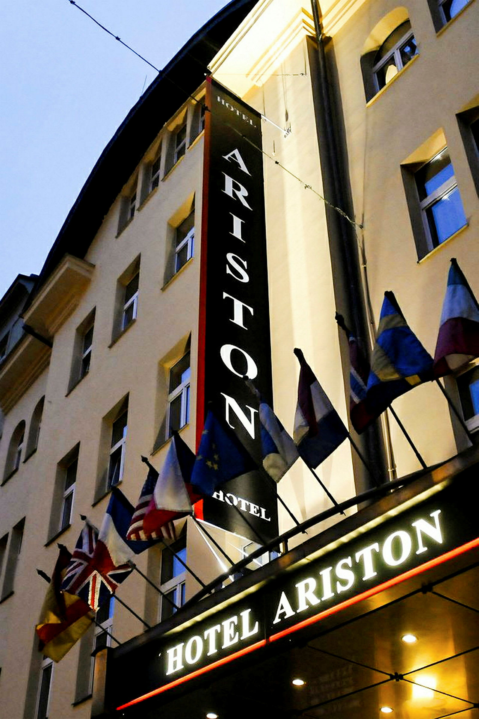 Hotellirakennus, jossa kyltissä lukee 'Hotel Ariston' ja sisäänkäynnin yläpuolella on useita kansallislippuja. (Tekoälyn luoma teksti)