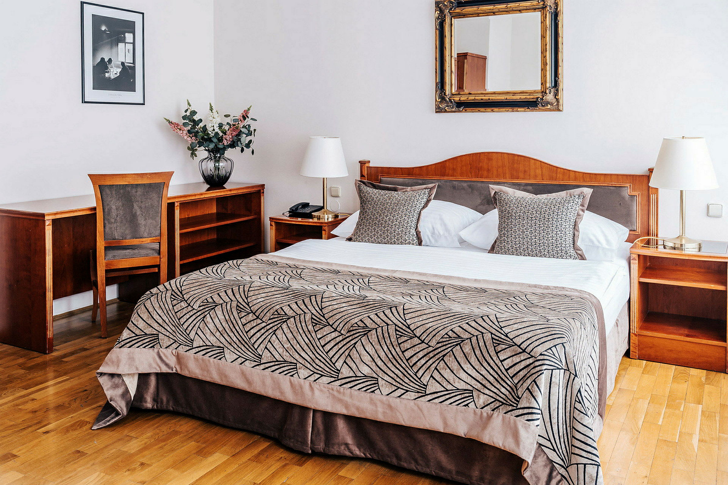Hotellihuone, jossa on parivuode, yöpöytä ja puinen kirjoituspöytä. Sängyllä on kuvioitu päiväpeitto ja tyynyjä. (Tekoälyn luoma teksti)