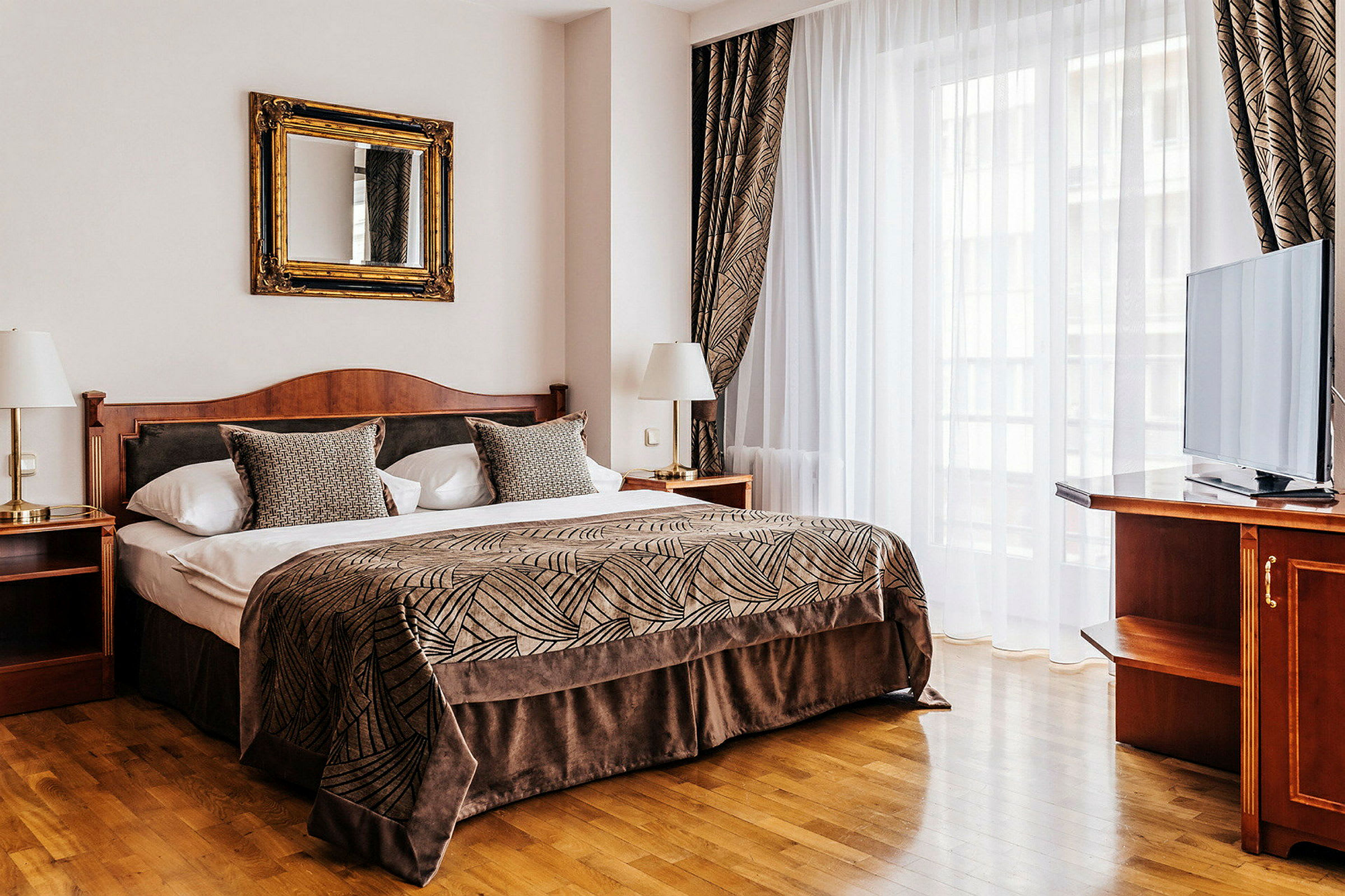 Tyylikäs hotellihuone, jossa on suuri sänky, verhoiltu kuvioidulla päiväpeitteellä ja koristeellisilla tyynyillä. Huoneessa on peili sängyn yläpuolella, kaksi yöpöytää lampuilla ja televisio huonekalulla suuren ikkunan vieressä. (Tekoälyn luoma teksti)