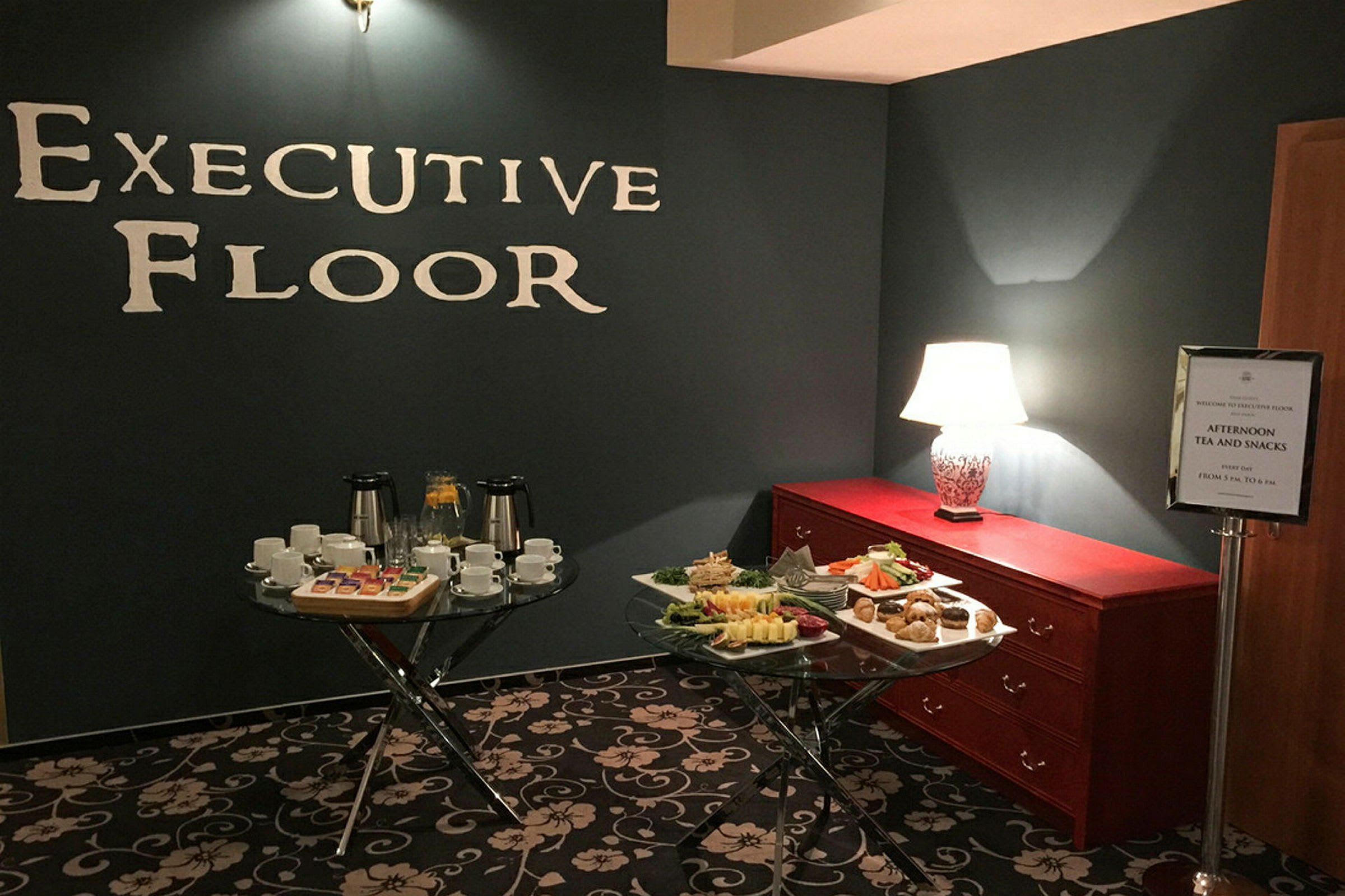 Huone, jossa seinällä on teksti "Executive Floor", moderni valaistus, kaksi pöytää kuppeineen, lautasineen ja naposteltavineen sekä lipasto pöytävalaisin. (Tekoälyn luoma teksti)