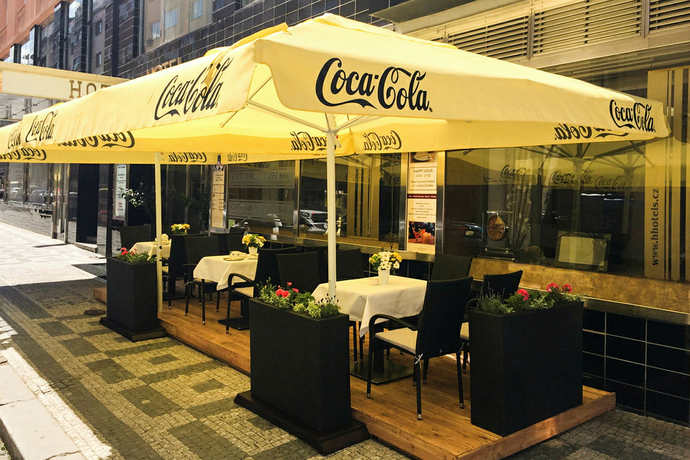 Ulkokahvila, jossa on pöytiä ja tuoleja suurten keltaisten, Coca-Cola-logolla merkittyjen aurinkovarjojen alla. (Tekoälyn luoma teksti)