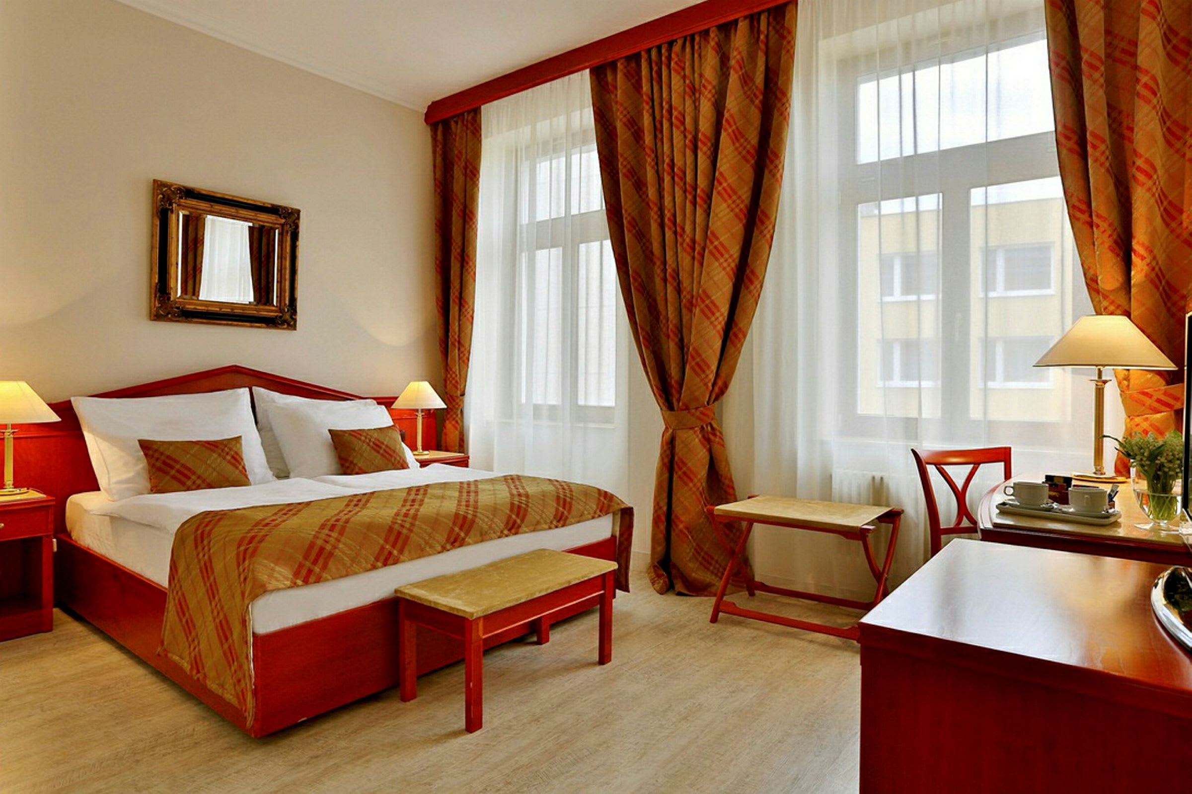 Hotellihuone, jossa on iso sänky ja kaksi lamppua yöpöydillä. Ikkunat verhoineen sekä pieni istumapaikka. (Tekoälyn luoma teksti)