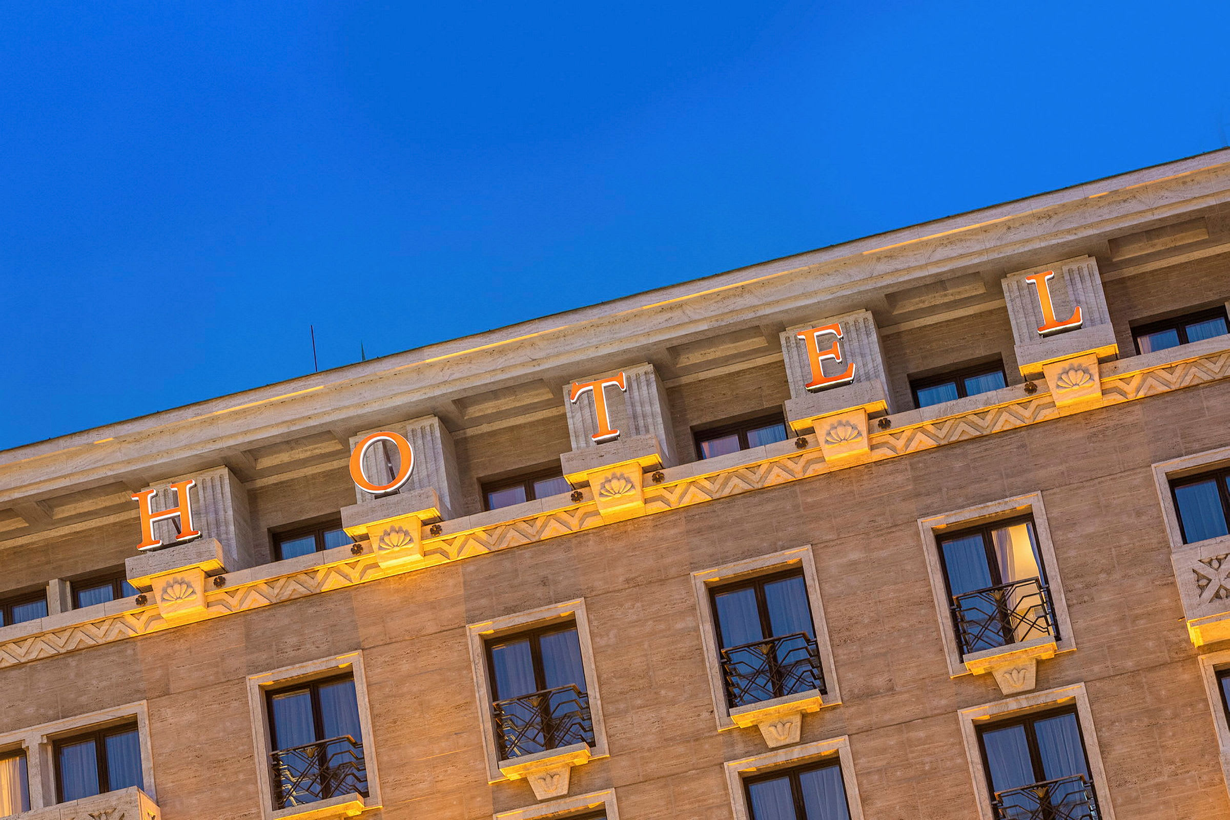 Hotellin julkisivu, jossa valaistut kirjaimet muodostavat sanan 'HOTEL' kirkasta sinistä iltataivasta vasten. (Tekoälyn luoma teksti)