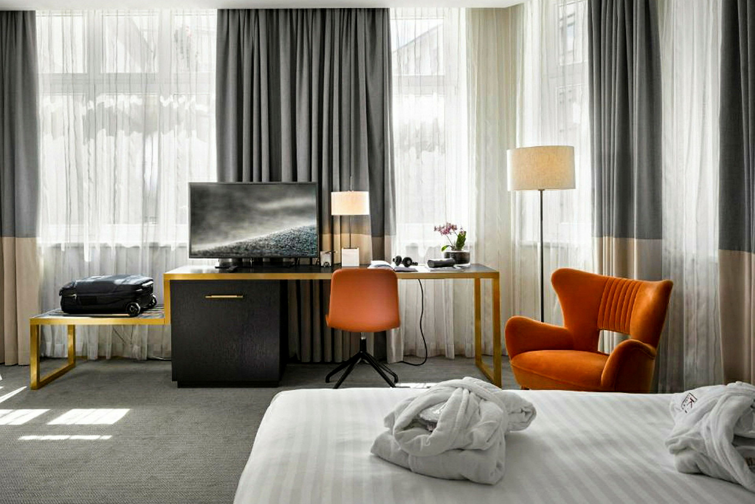 Hotellihuone, jossa etualalla on sänky valkoisilla vuodevaatteilla, ja taustalla istuinryhmä, jossa on oranssi tuoli ja työpöytä televisiolla. (Tekoälyn luoma teksti)