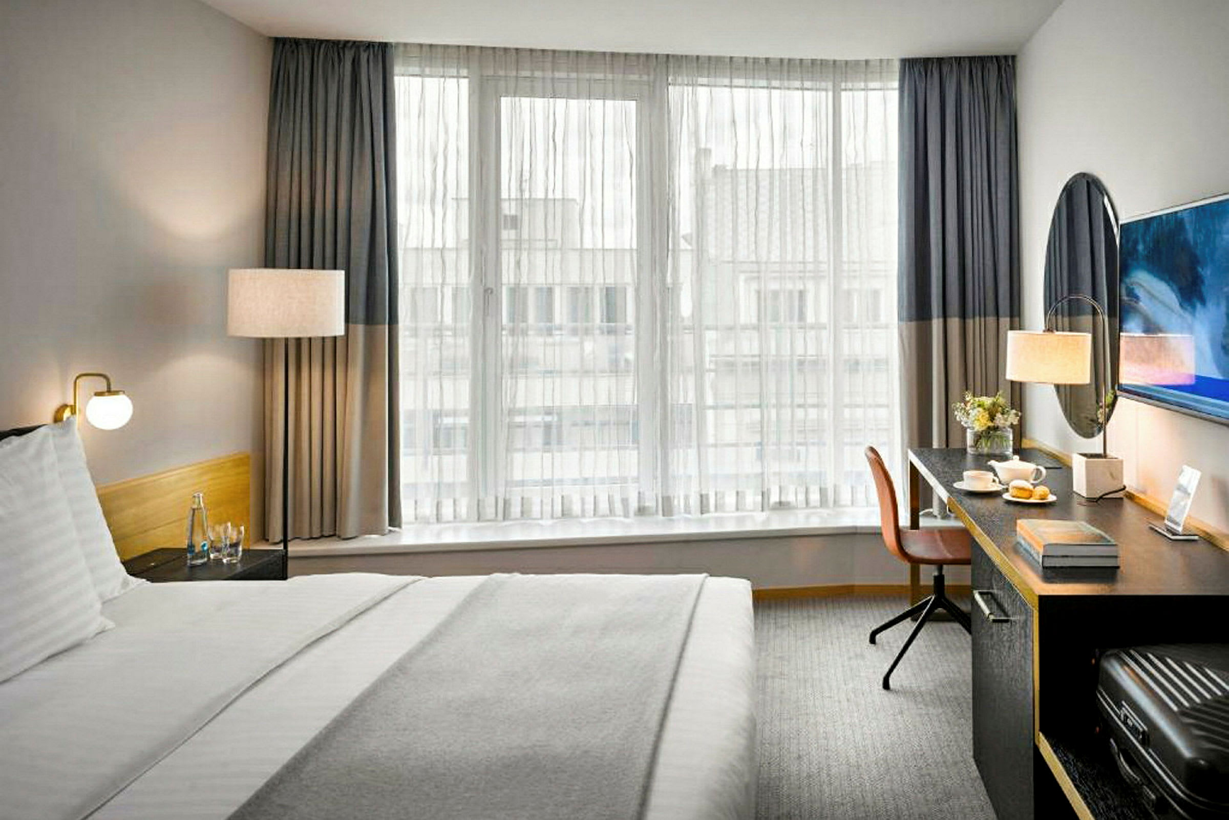 Hotellihuone, jossa on sänky, kirjoituspöytä ja ikkuna ohuilla verhoilla. (Tekoälyn luoma teksti)