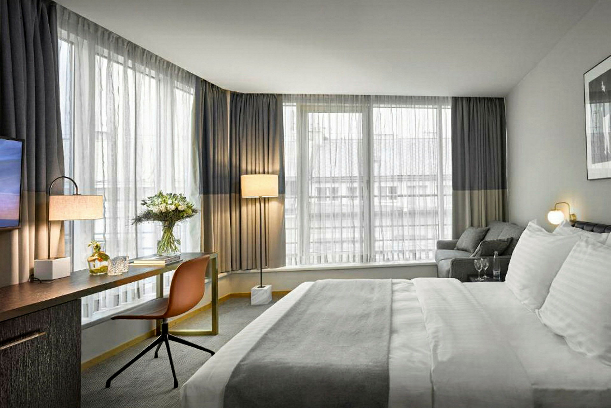 Hotellihuone, jossa suuri sänky, kirjoituspöytä, tuoli ja ikkunat verhoilla. (Tekoälyn luoma teksti)