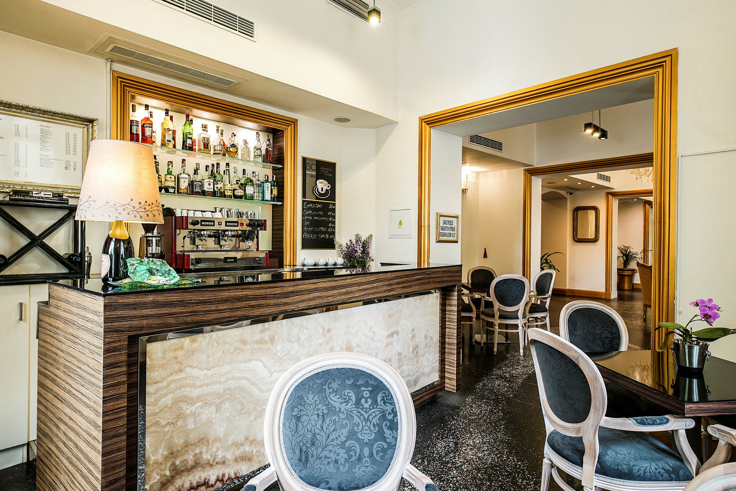 Kuva pienestä hotellibaarista, jossa on erilaisia alkoholijuomia hyllyllä sekä kahvikone, kalusteiden ympäröimänä. (Tekoälyn luoma teksti)