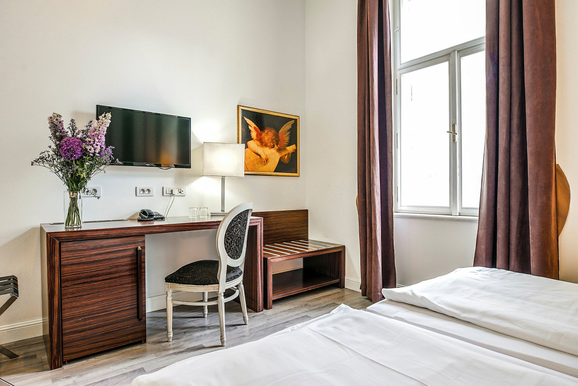Hotellihuone, jossa on kirjoituspöytä ja tuoli, seinään kiinnitetty televisio, kukkakimppu ja lamppu pöydällä sekä suuri ikkuna ruskeilla verhoilla. (Tekoälyn luoma teksti)