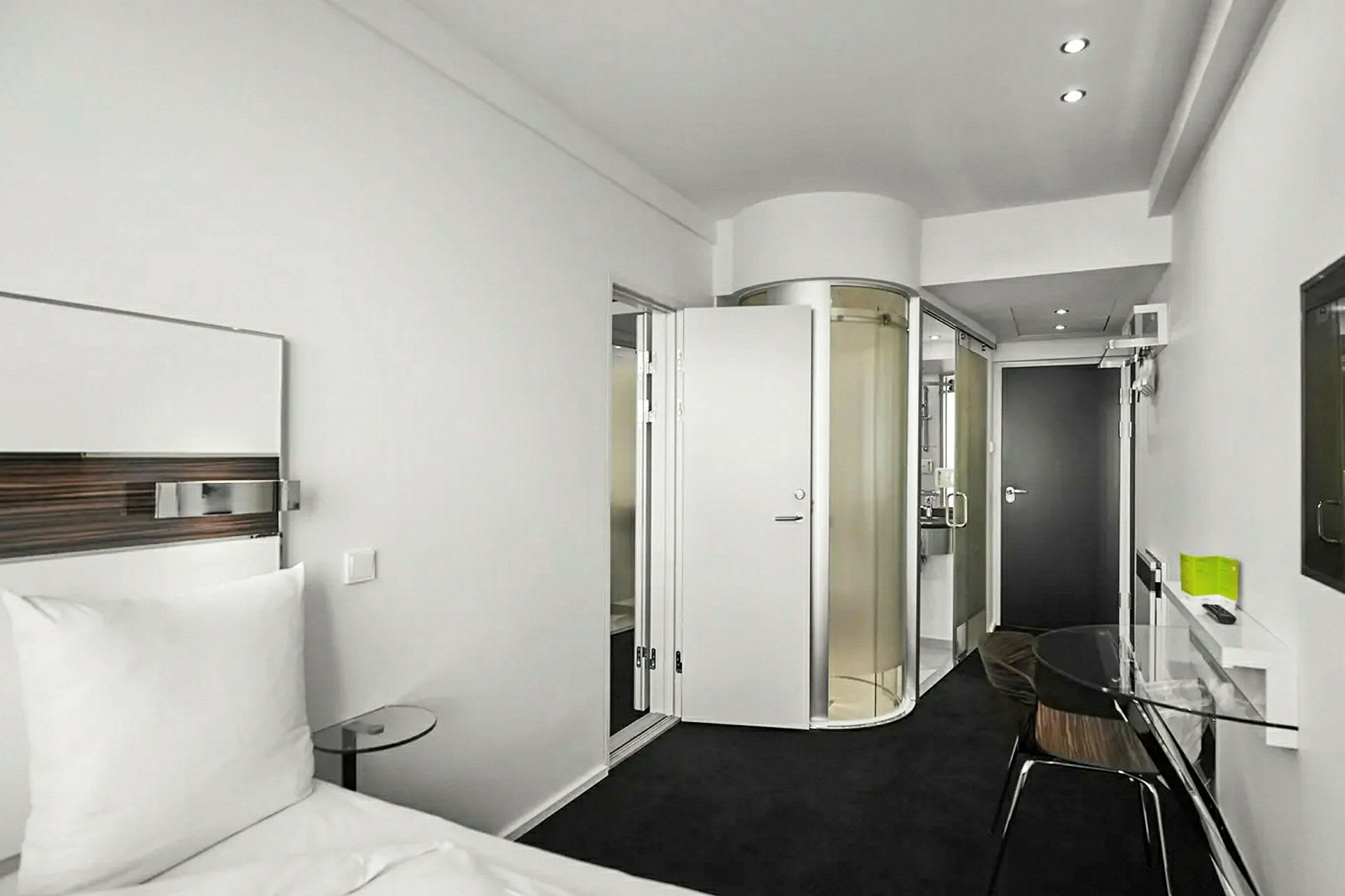 Hotellihuone, jossa on moderni sisustus. Vuode valkoisilla liinavaatteilla, lasipöytä ja pyöreä suihkukaappi näkyvät. (Tekoälyn luoma teksti)