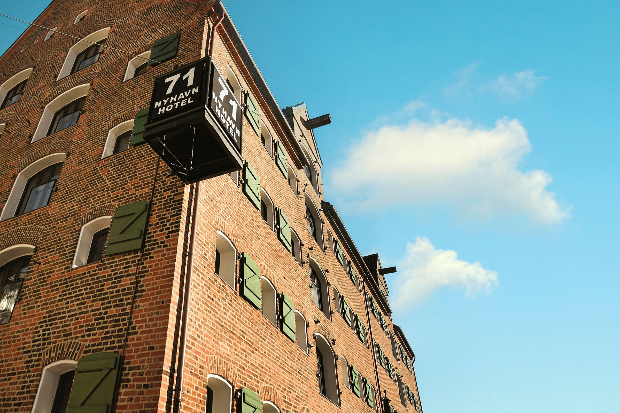 Tiilirakennus, jossa on vihreät ikkunaluukut ja kyltti, jossa lukee "71 NYHAVN HOTEL" sinistä taivasta vasten. (Tekoälyn luoma teksti)