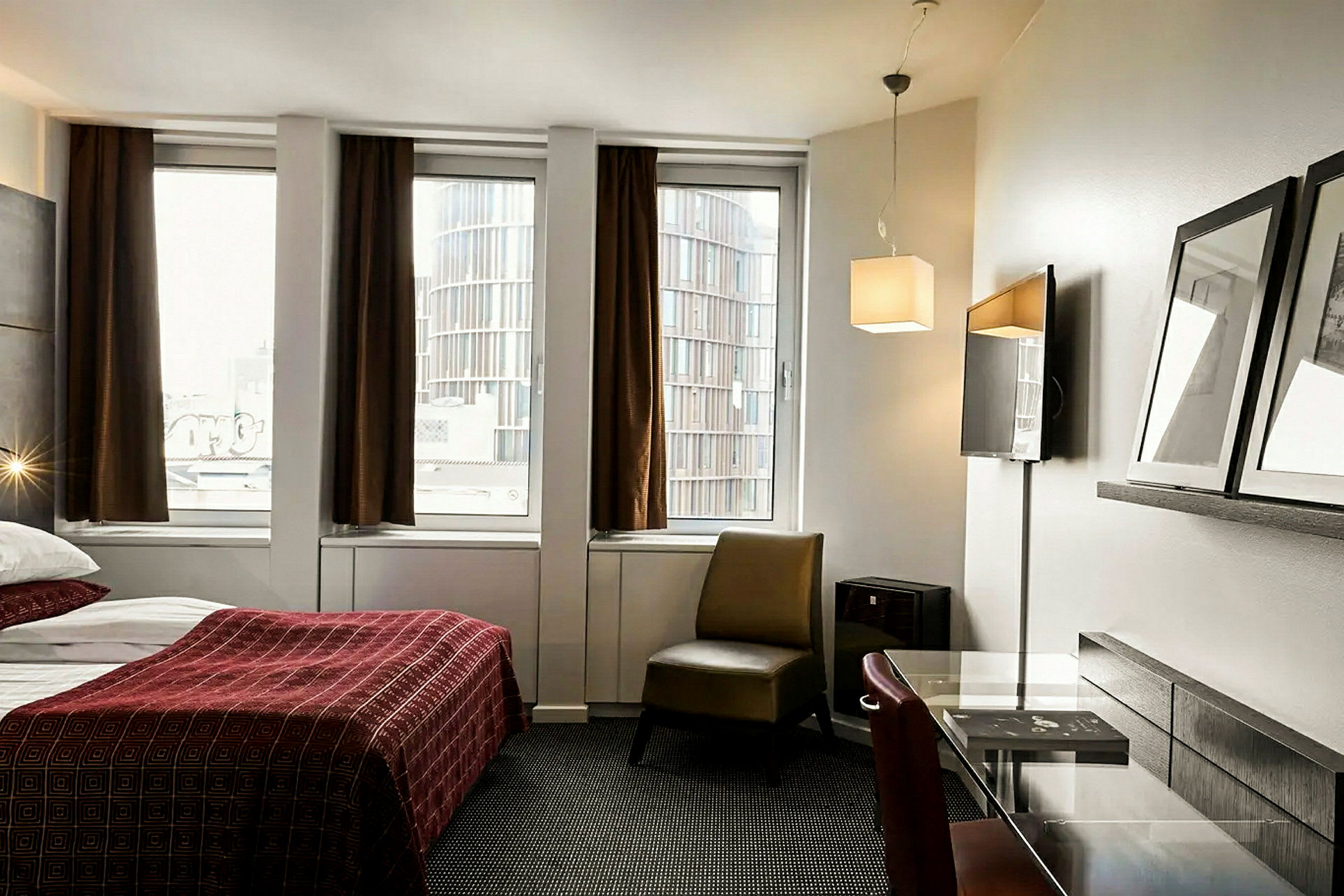 Hotellihuone, jossa on punaisella päiväpeitteellä varustettu sänky, työpöytä ja tuoli kolmen ikkunan edessä, joissa ruskeat verhot. (Tekoälyn luoma teksti)