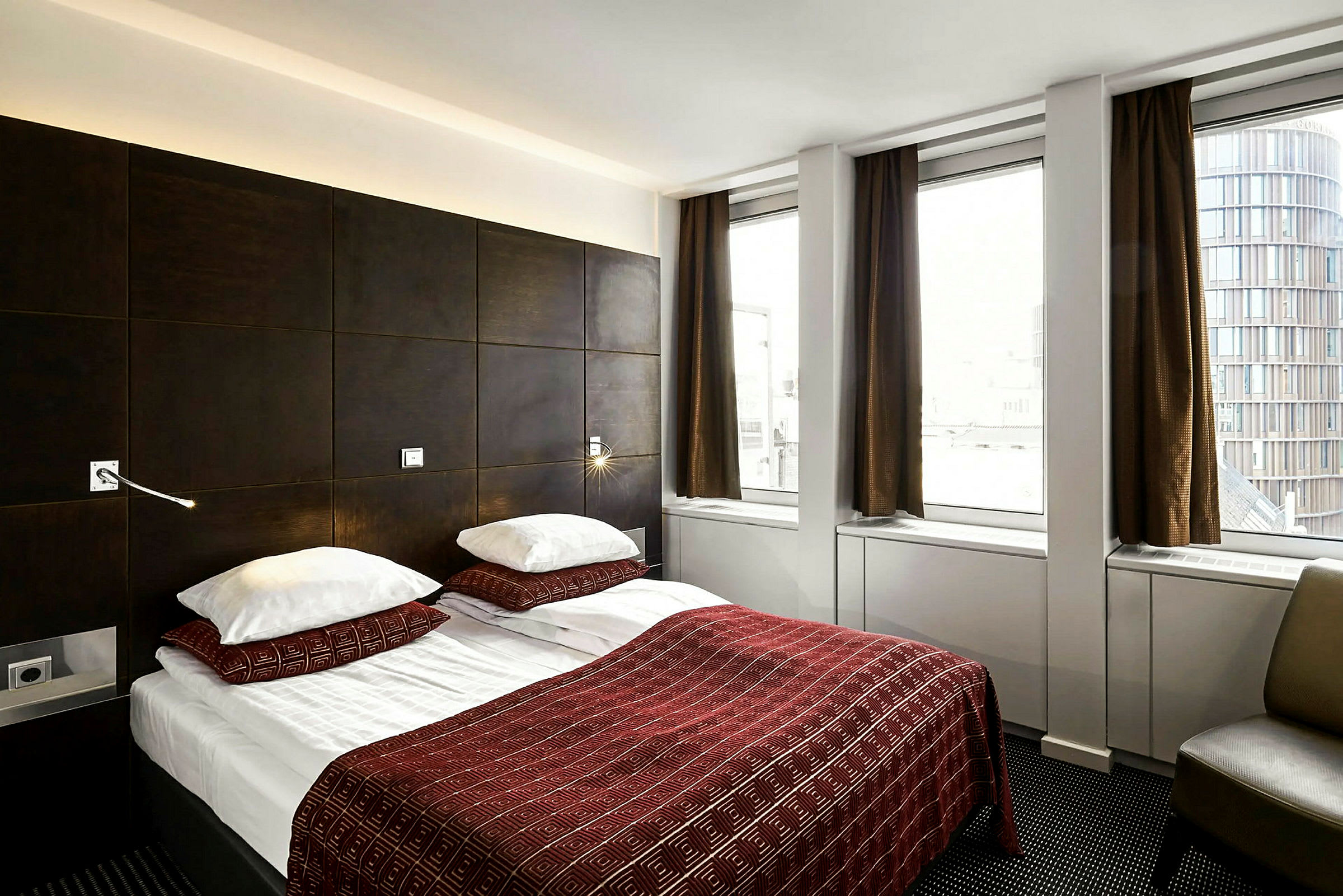 Hotellihuone, jossa on parivuode, peitetty punaisella kuvioidulla päiväpeitteellä ja kahdella tyynyllä. Huoneessa on tummat huonekalut ja kolme suurta ikkunaa ruskeilla verhoilla. (Tekoälyn luoma teksti)