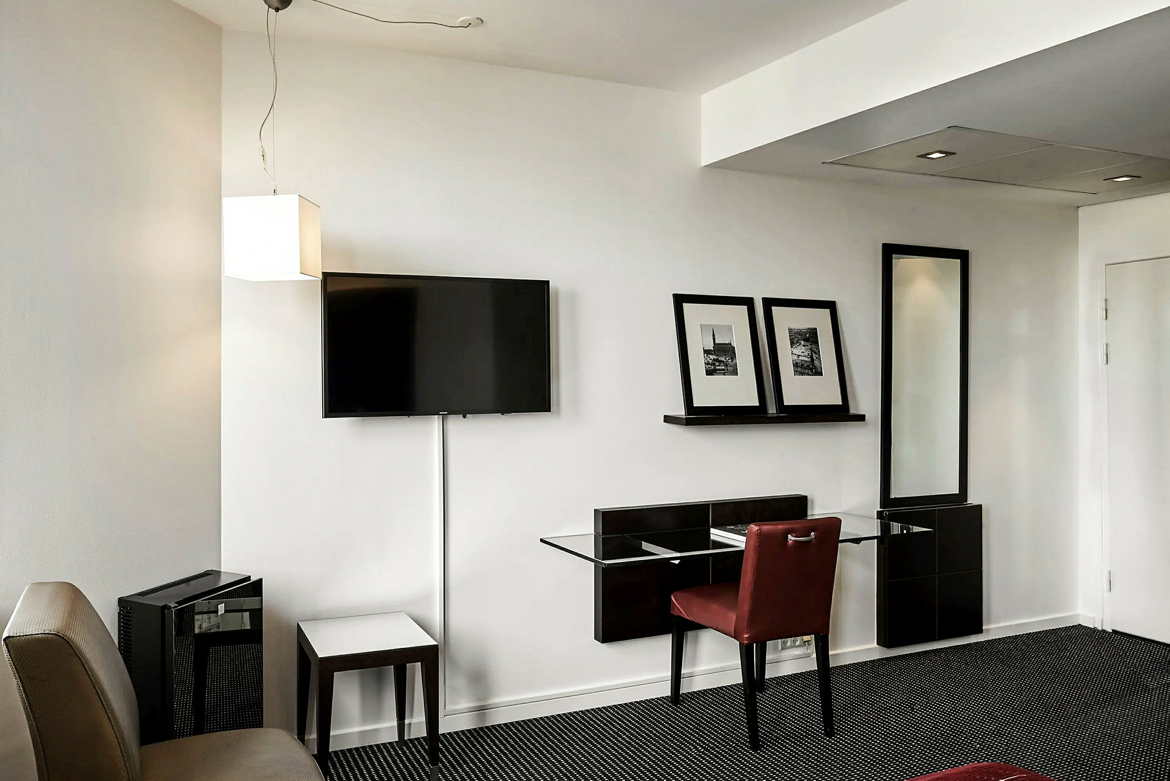 Hotellihuone, jossa seinällä on seinälle asennettu televisio, hylly kahdella kehystetyllä kuvalla ja istuinalue pöytineen ja tuoleineen. (Tekoälyn luoma teksti)
